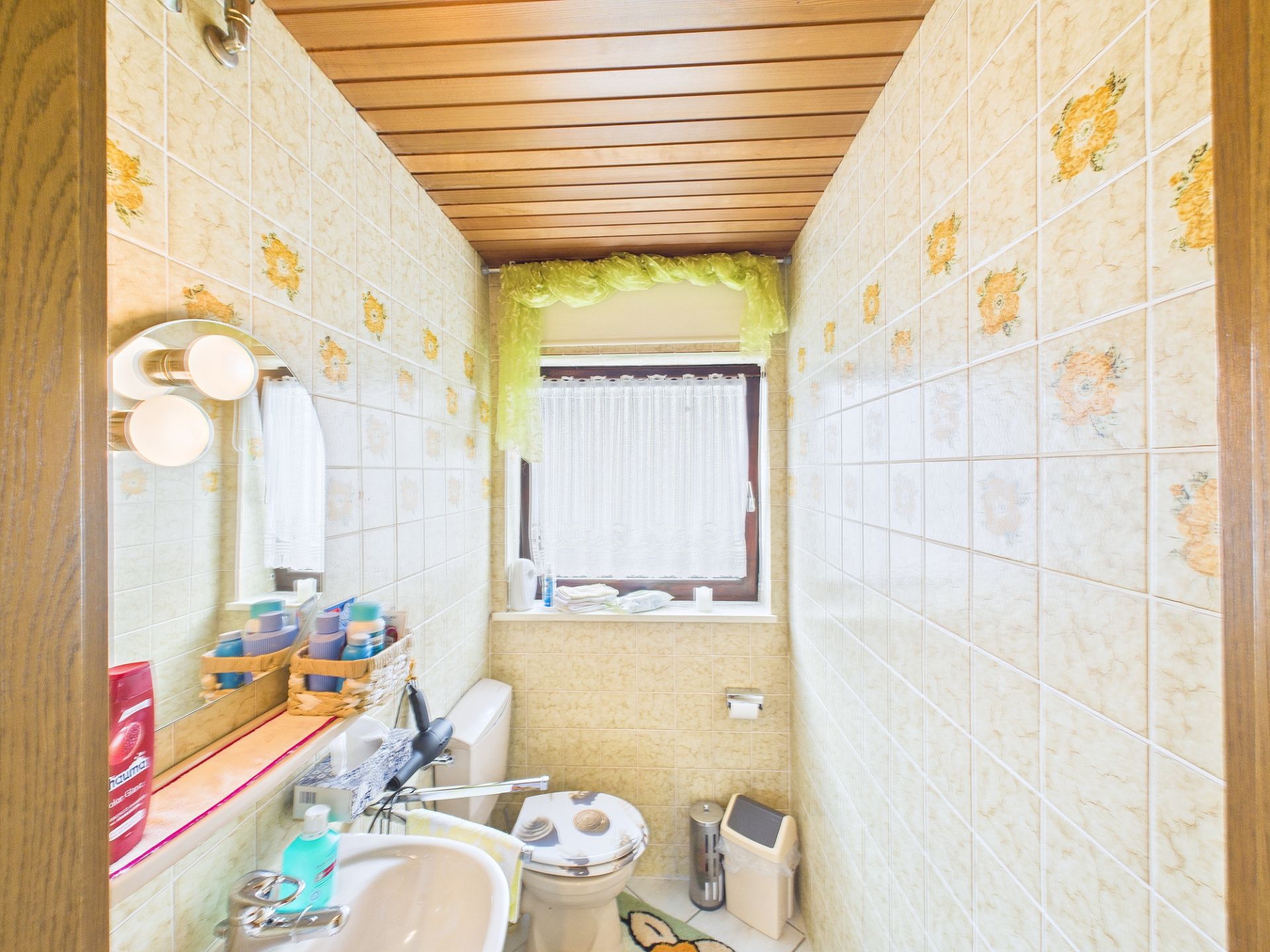 Kleines Bad mit WC, Waschbecken, Blumenfliesen und Fenster mit Gardine.