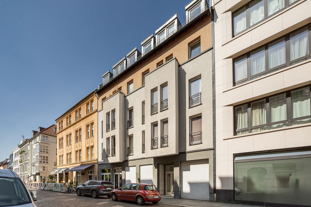 Mehrstöckiges Wohnhaus mit Balkonen und geparkten Autos in einer Stadtstraße bei sonnigem Wetter.