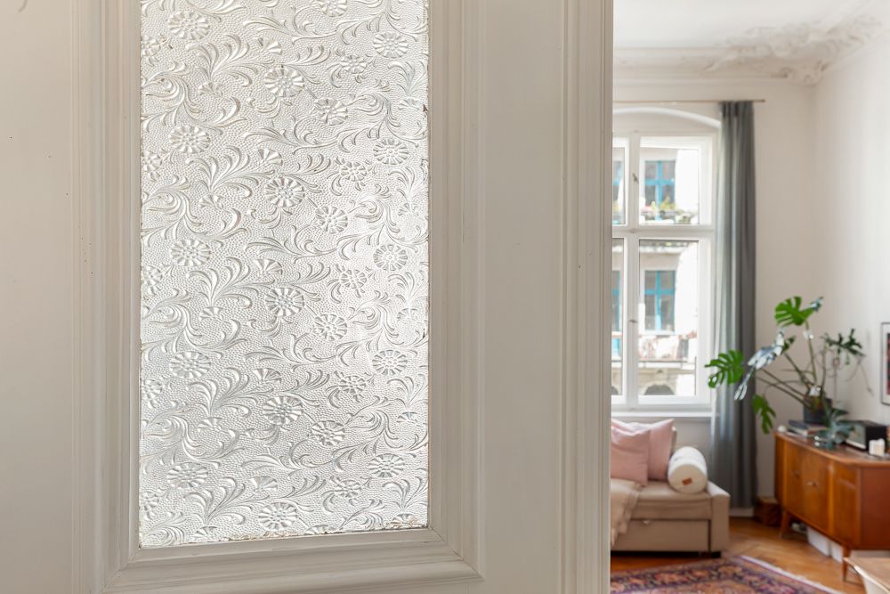 Weiße Tür mit Milchglas und Blumenmuster, dahinter ein Wohnzimmer mit Sofa und Pflanzen.