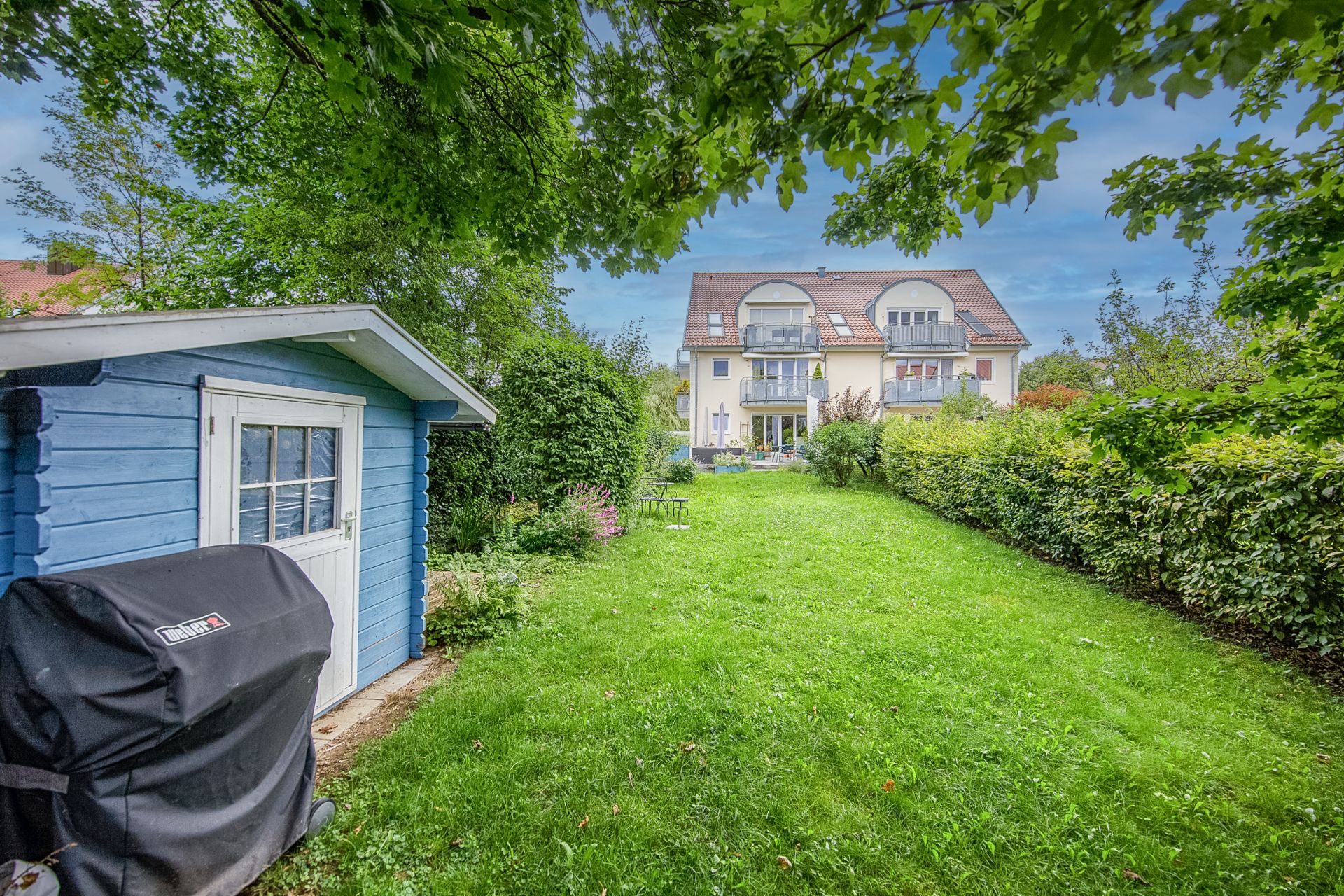 Grüner Garten mit blauem Gartenschuppen und Haus im Hintergrund.