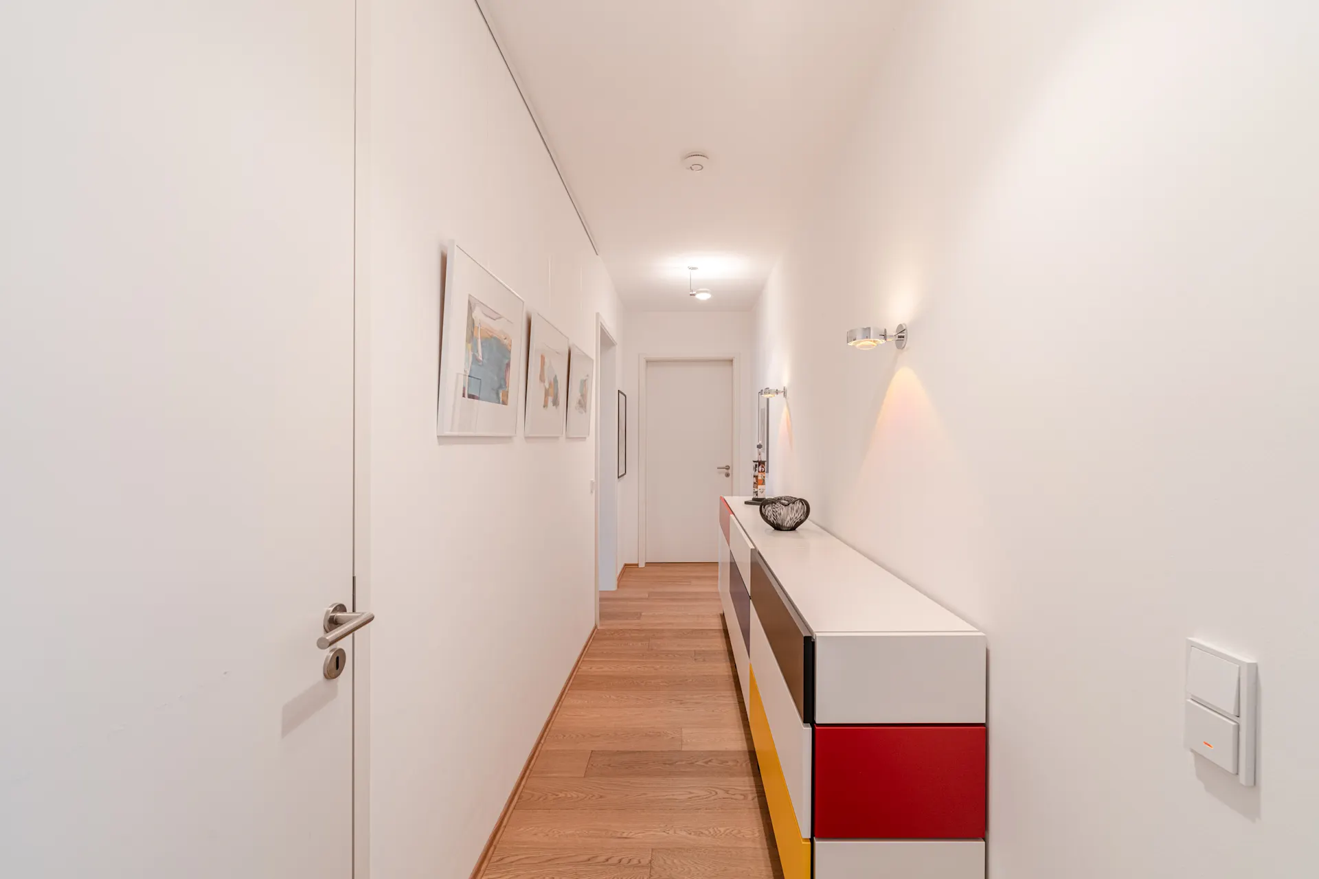 Heller Flur mit Bildern an der Wand und bunter Kommode, Holzfußboden.