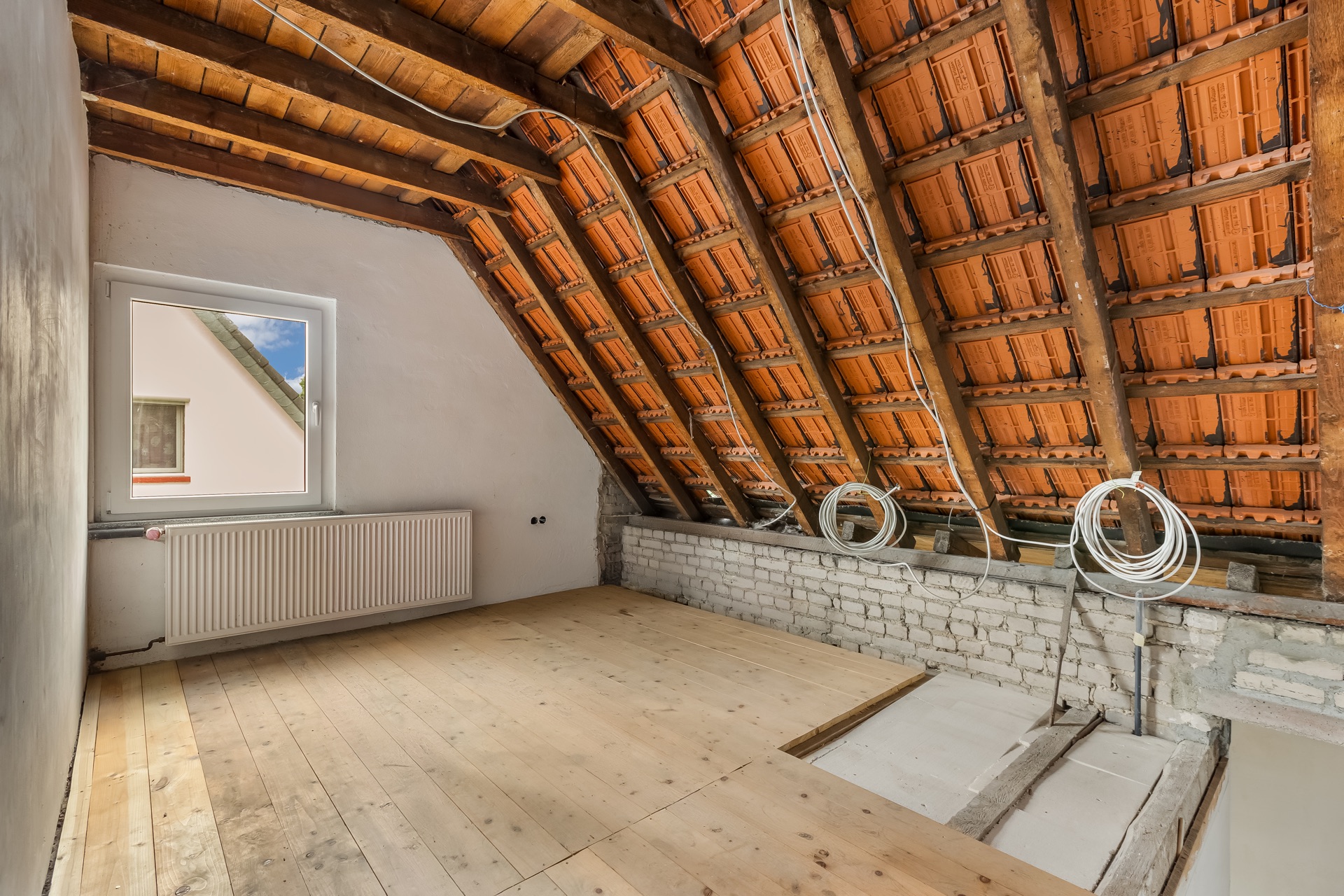 Dachbodenraum mit sichtbaren Holzbalken, Ziegeln, Holzfußboden und einem Fenster.