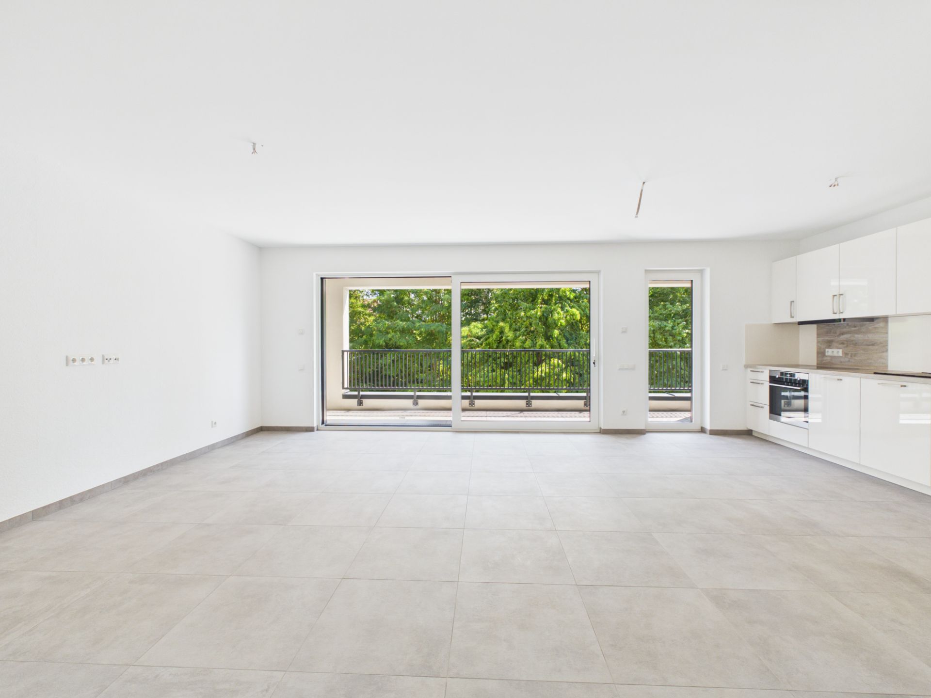 Heller, leerer Raum mit Fliesenboden, Einbauküche und großen Fenstern zu einem Balkon.
