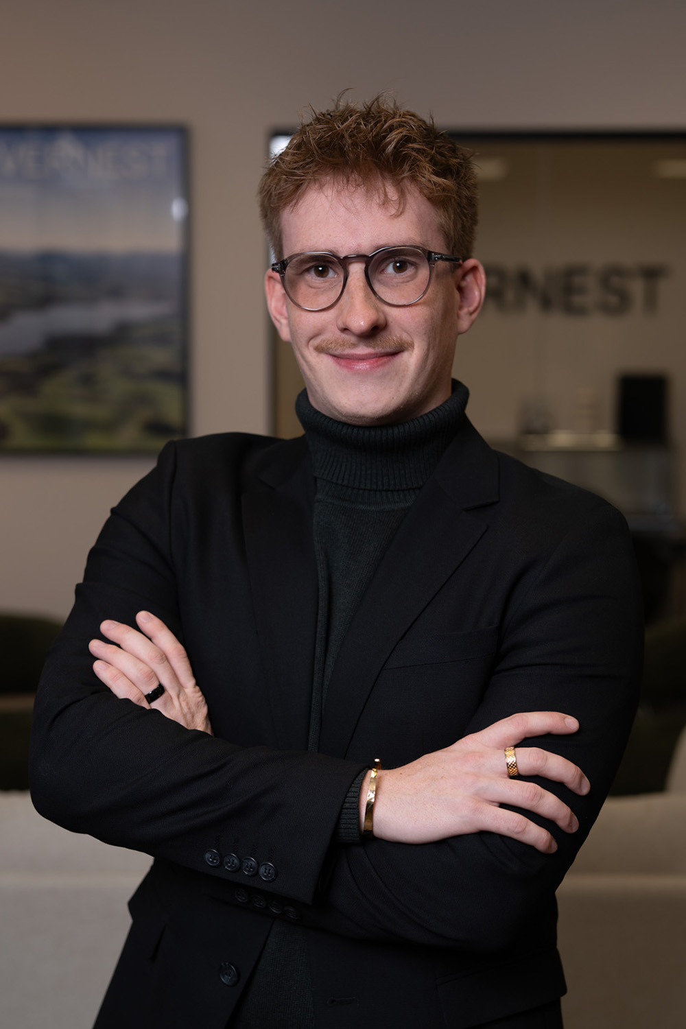 Junger Mann mit Brille und kariertem Sakko steht in modernem Innenraum.