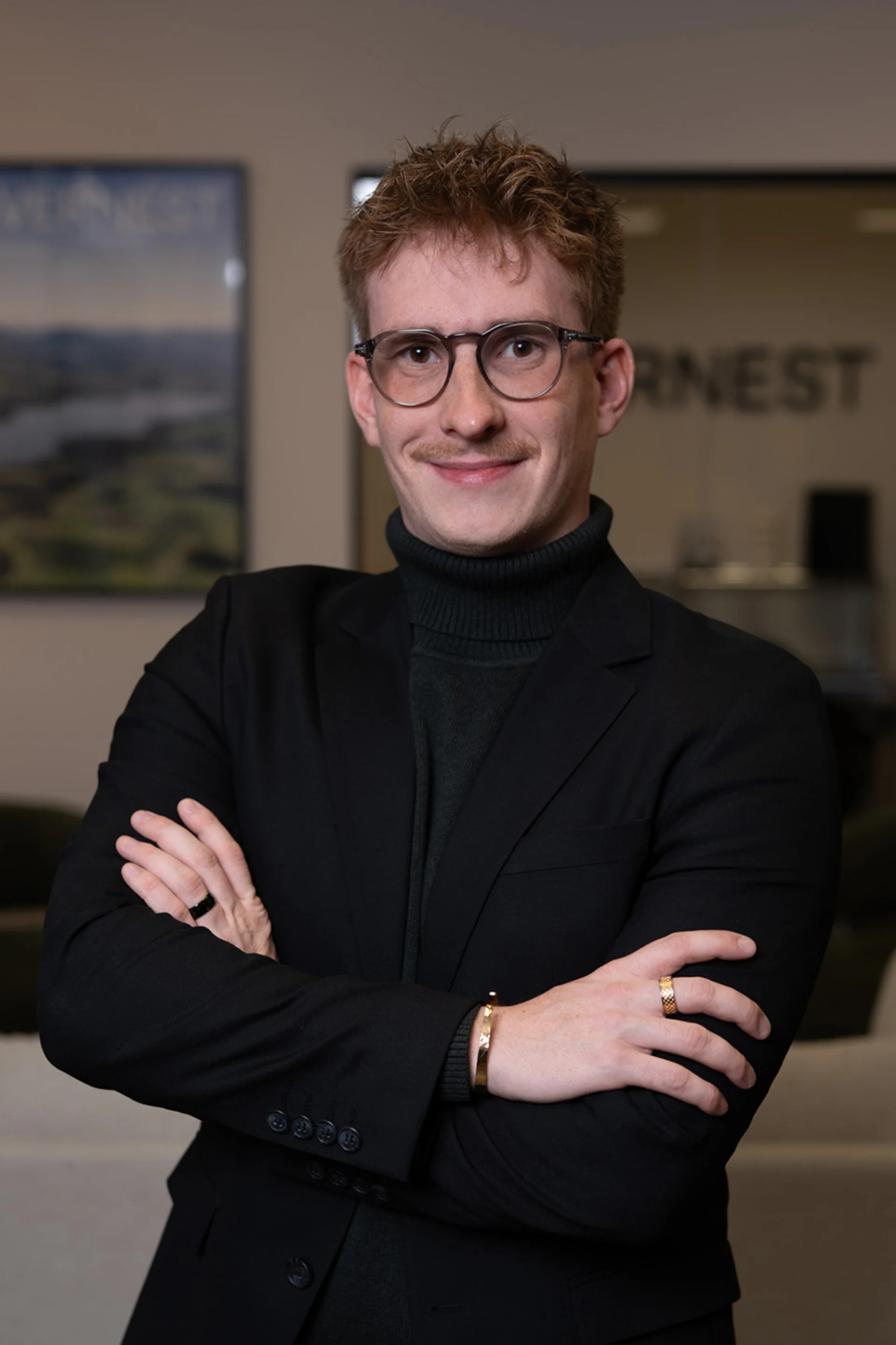 Junger Mann mit Brille und kariertem Sakko steht in modernem Innenraum.