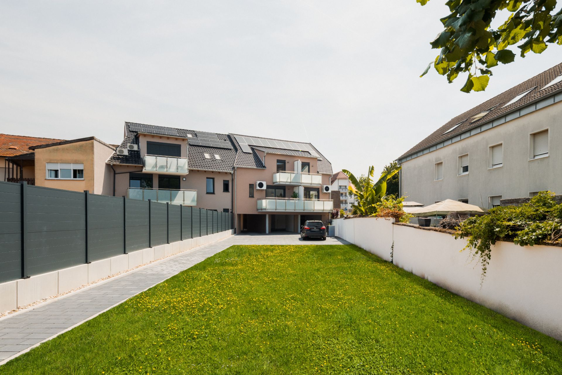 Mehrfamilienhaus mit Balkonen, davor eine Einfahrt, Rasenfläche und Gartenzaun.