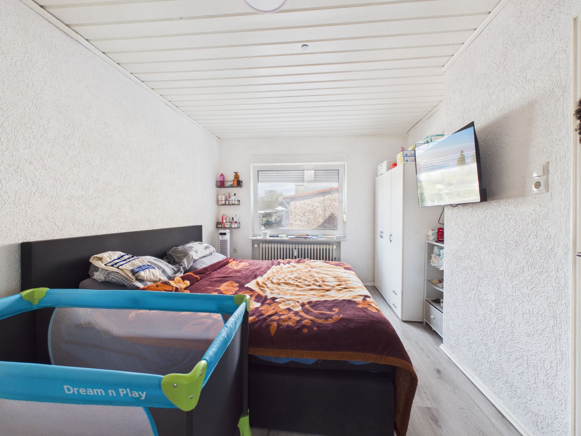 Schlafzimmer mit Doppelbett, Babybett, Schrank und Wandfernseher, Fenster an der Stirnwand.