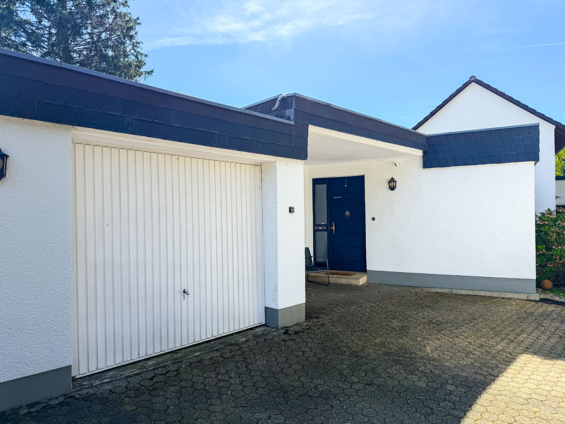 Weiße Hausfassade mit Garage und blauem Eingang, davor gepflasterter Hof.