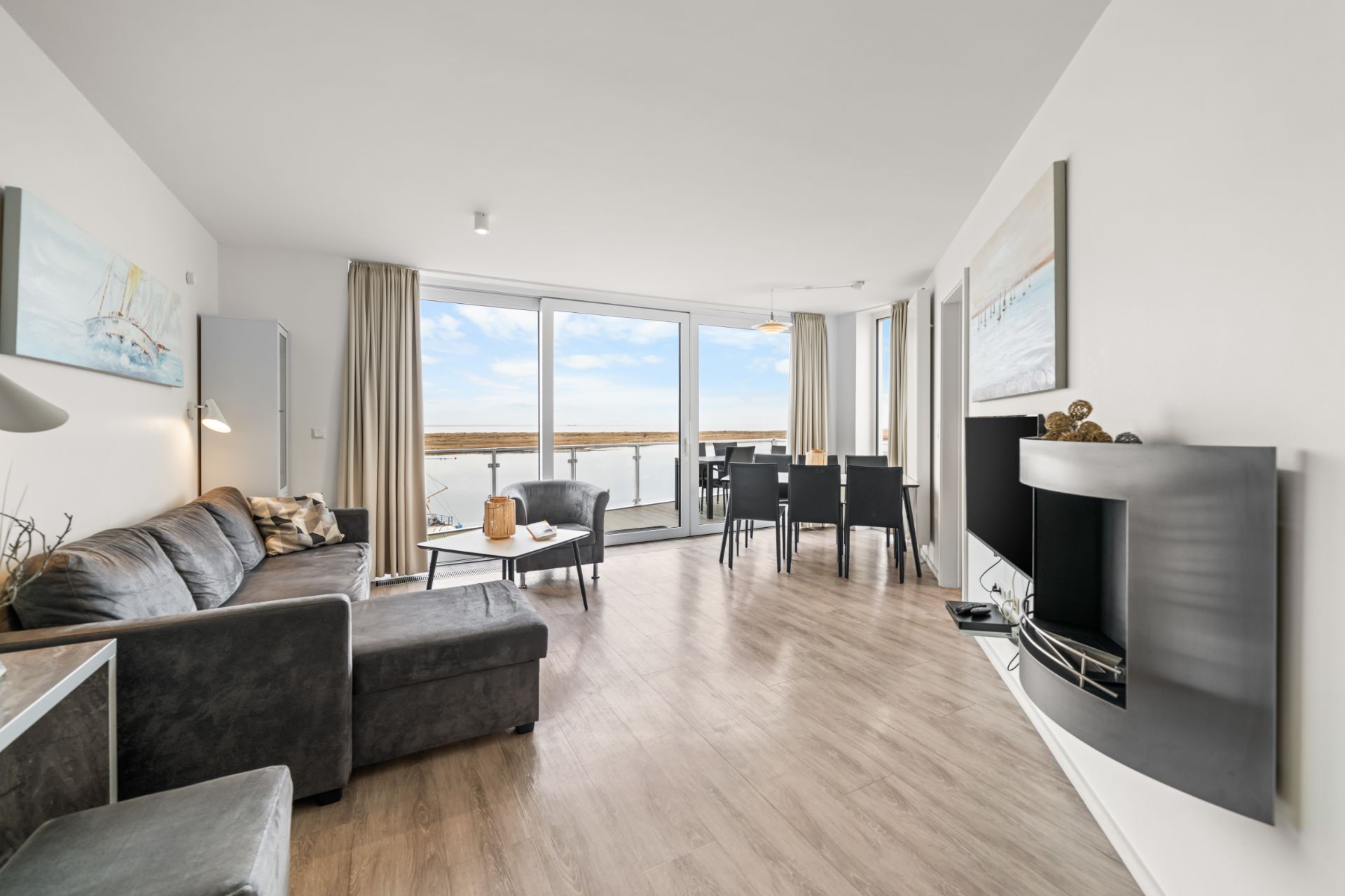 Helles Wohnzimmer mit Sofa, Esstisch, großem Fenster und Blick auf Balkon und Wasser.