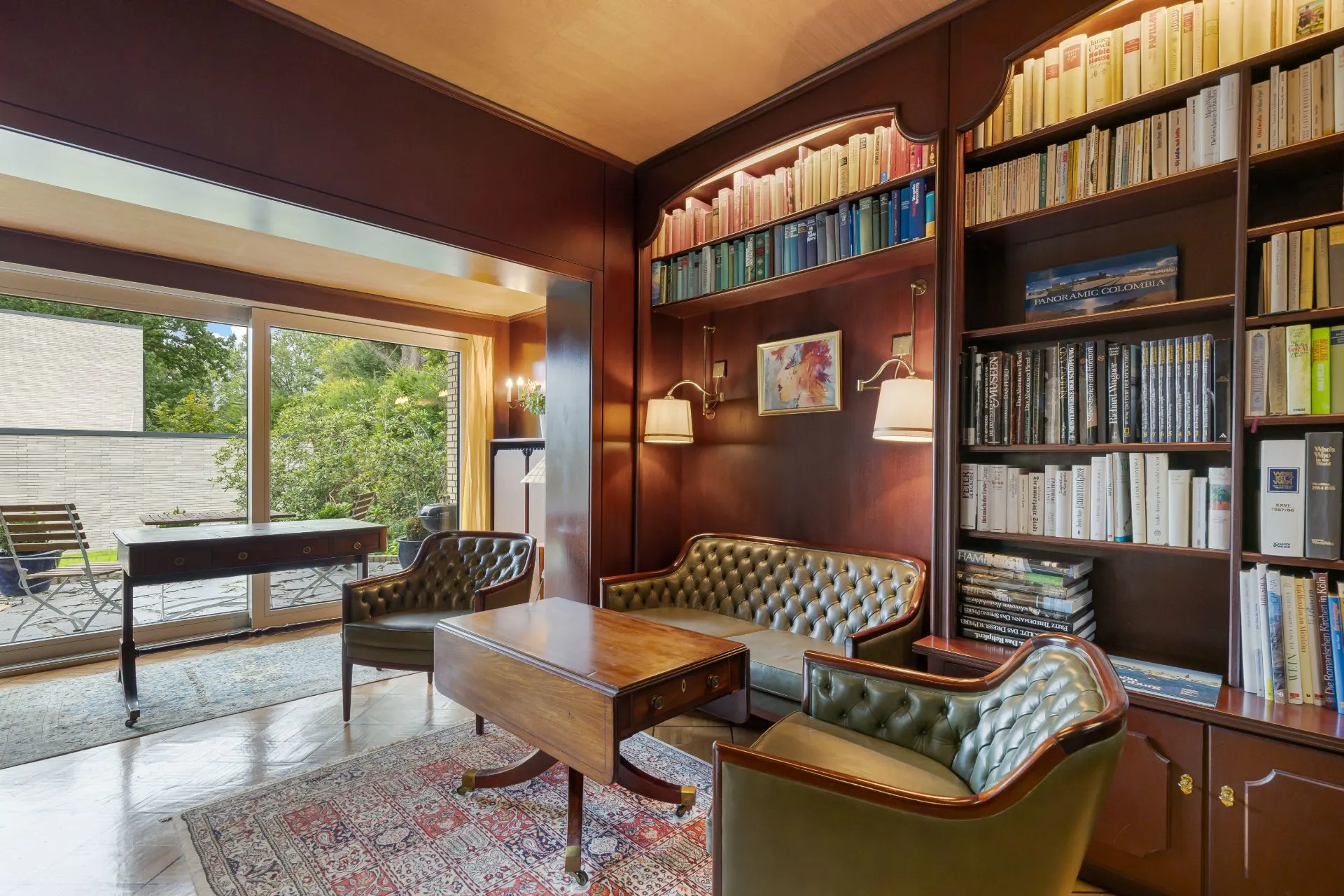 Gemütliches Wohnzimmer mit Bücherregalen, Lederstühlen, Sofa, Holztisch und Blick auf Terrasse mit Garten.