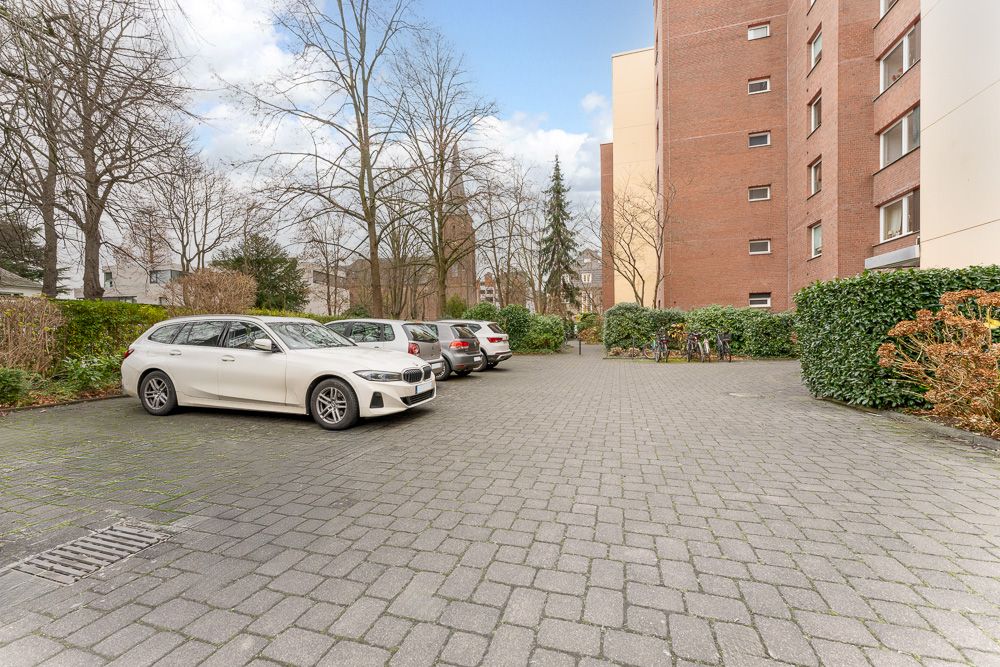 Parkplatz mit mehreren Autos neben einem Wohnhaus, umgeben von Bäumen und Sträuchern.