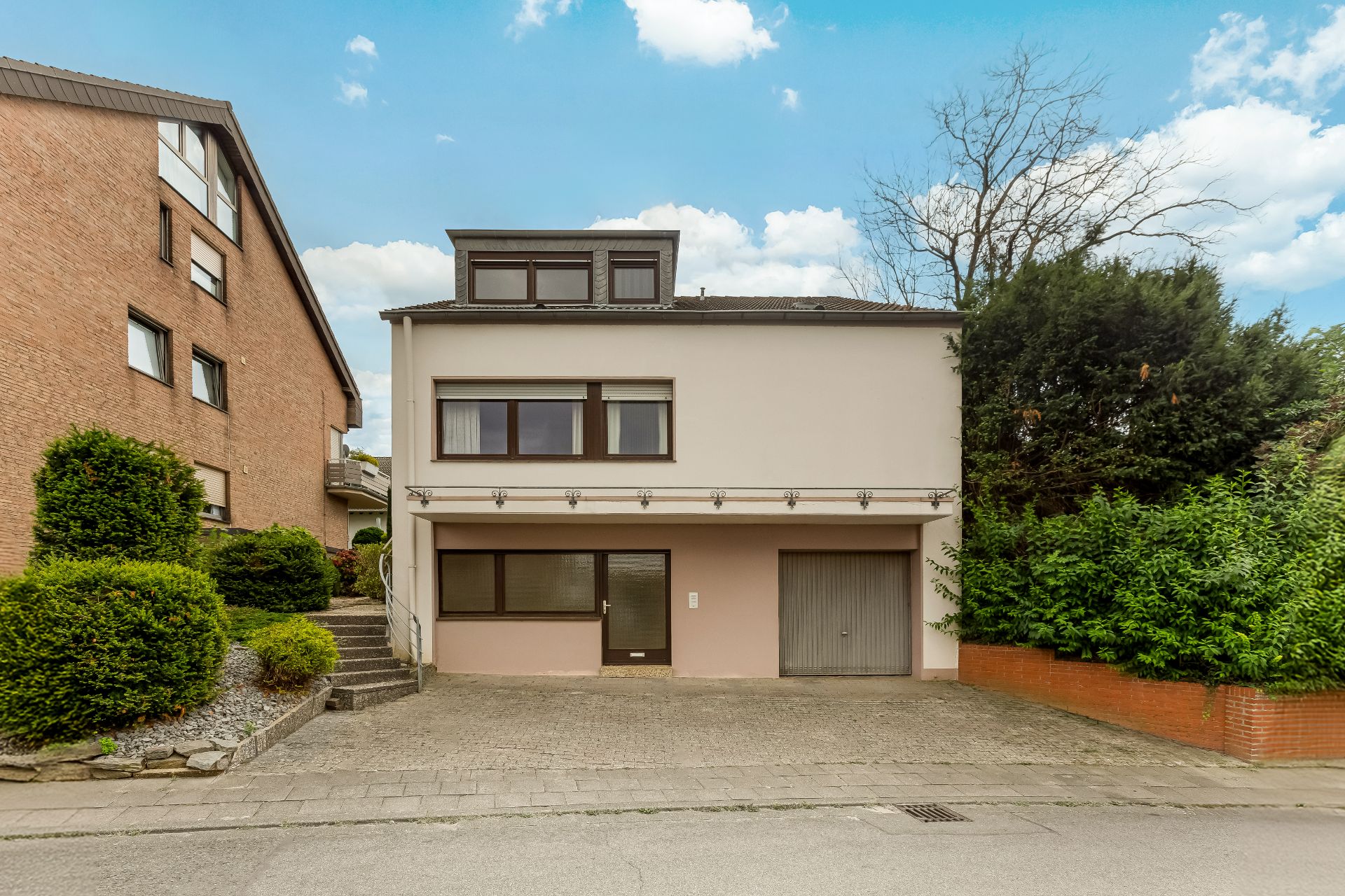 Vorderansicht eines hellen Wohnhauses mit Garage, großen Fenstern und gepflasterter Einfahrt.