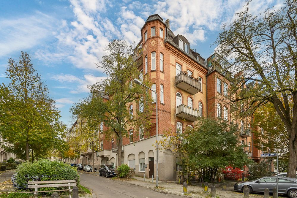 Mehrstöckiges rotes Wohnhaus an einer Straßenecke mit Balkonen und Bäumen davor.