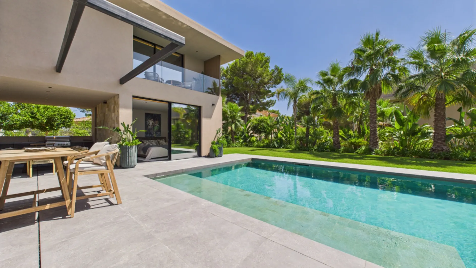 Moderne Terrasse eines Hauses mit Pool, Garten und Palmen bei sonnigem Wetter.