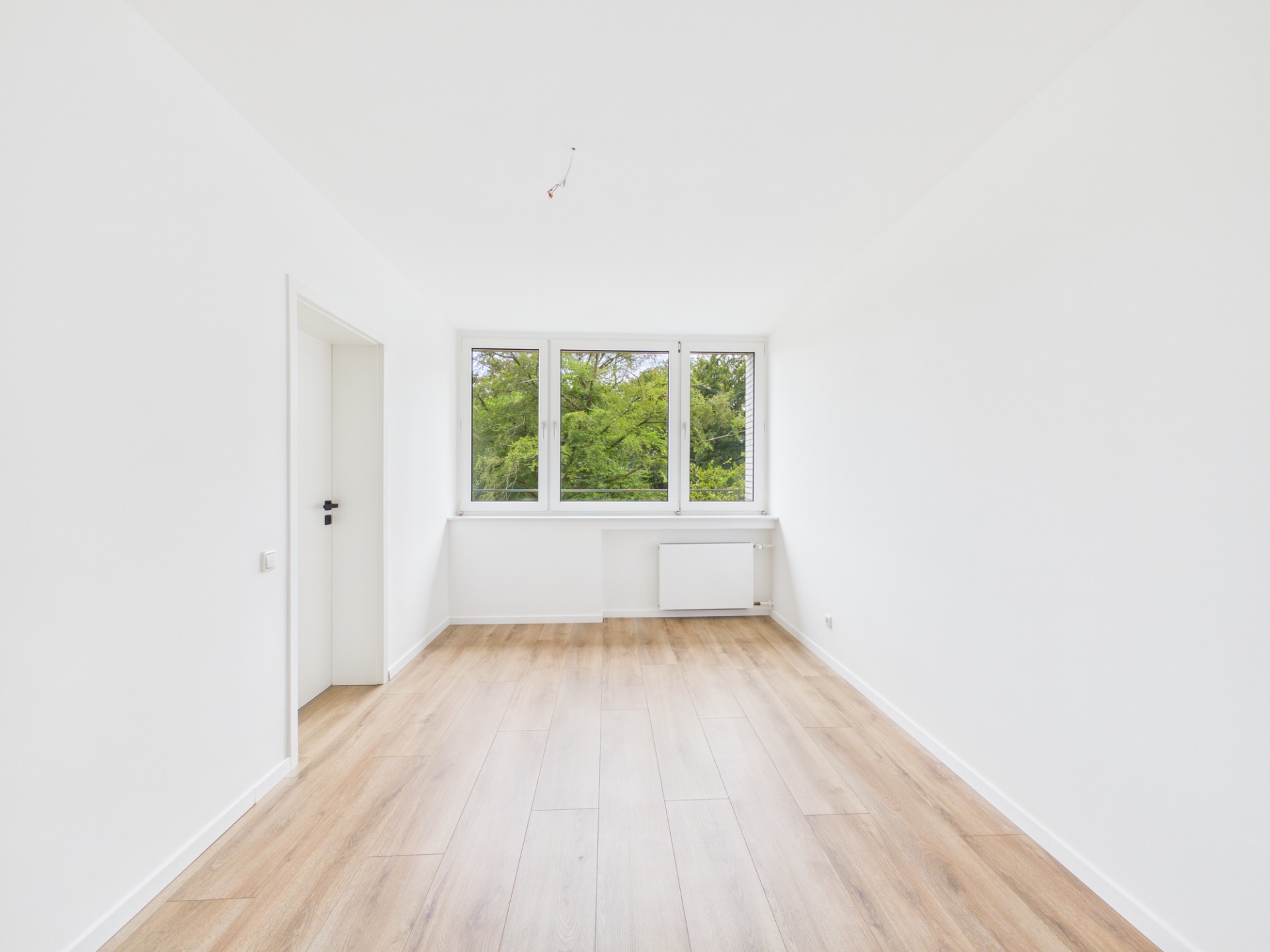 Heller, leerer Raum mit Holzfußboden, weißen Wänden und großem Fenster mit Blick ins Grüne.