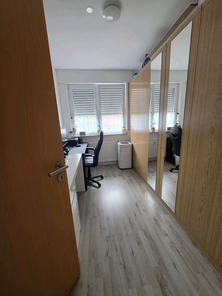 Helles Arbeitszimmer mit Schreibtisch, Stuhl, Spiegelschrank und drei Fenstern.