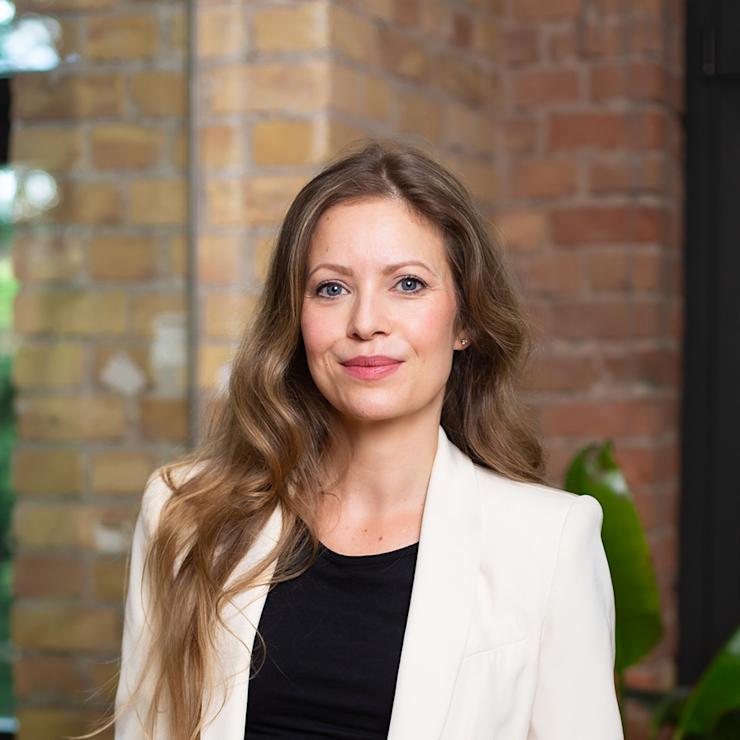 Frau mit langen Haaren und weißem Blazer steht vor einer Backsteinwand im Innenraum.