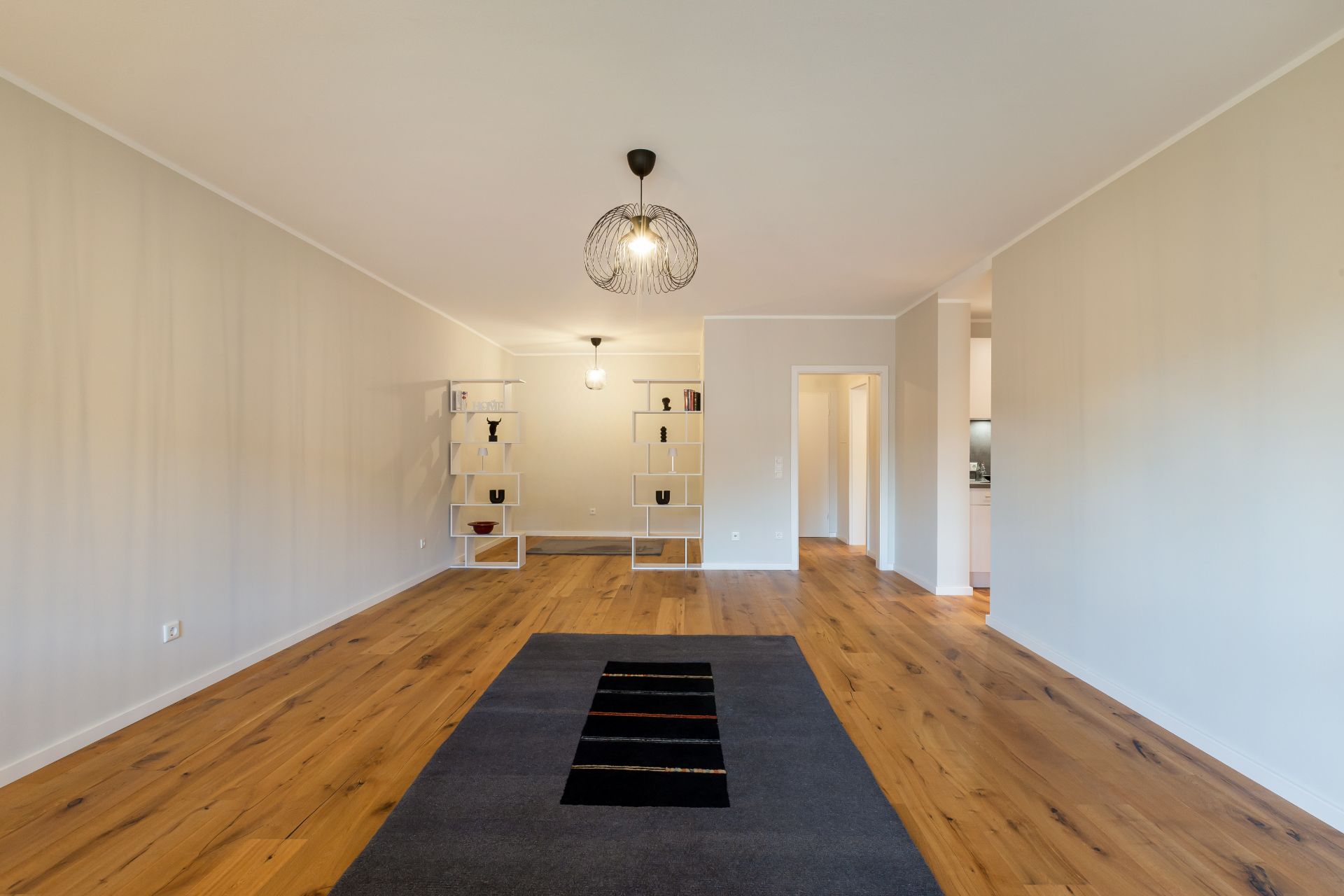 Helles Wohnzimmer mit Holzboden, Teppich, Regal und moderner Deckenleuchte.
