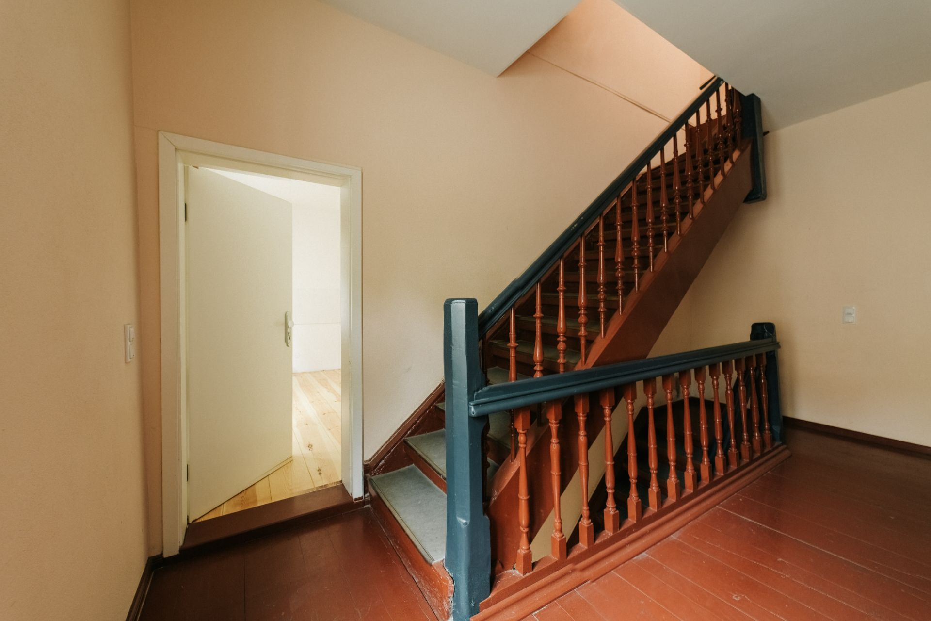 Ein Treppenhaus mit brauner Holztreppe und offen stehender Tür zu einem hellen Raum.