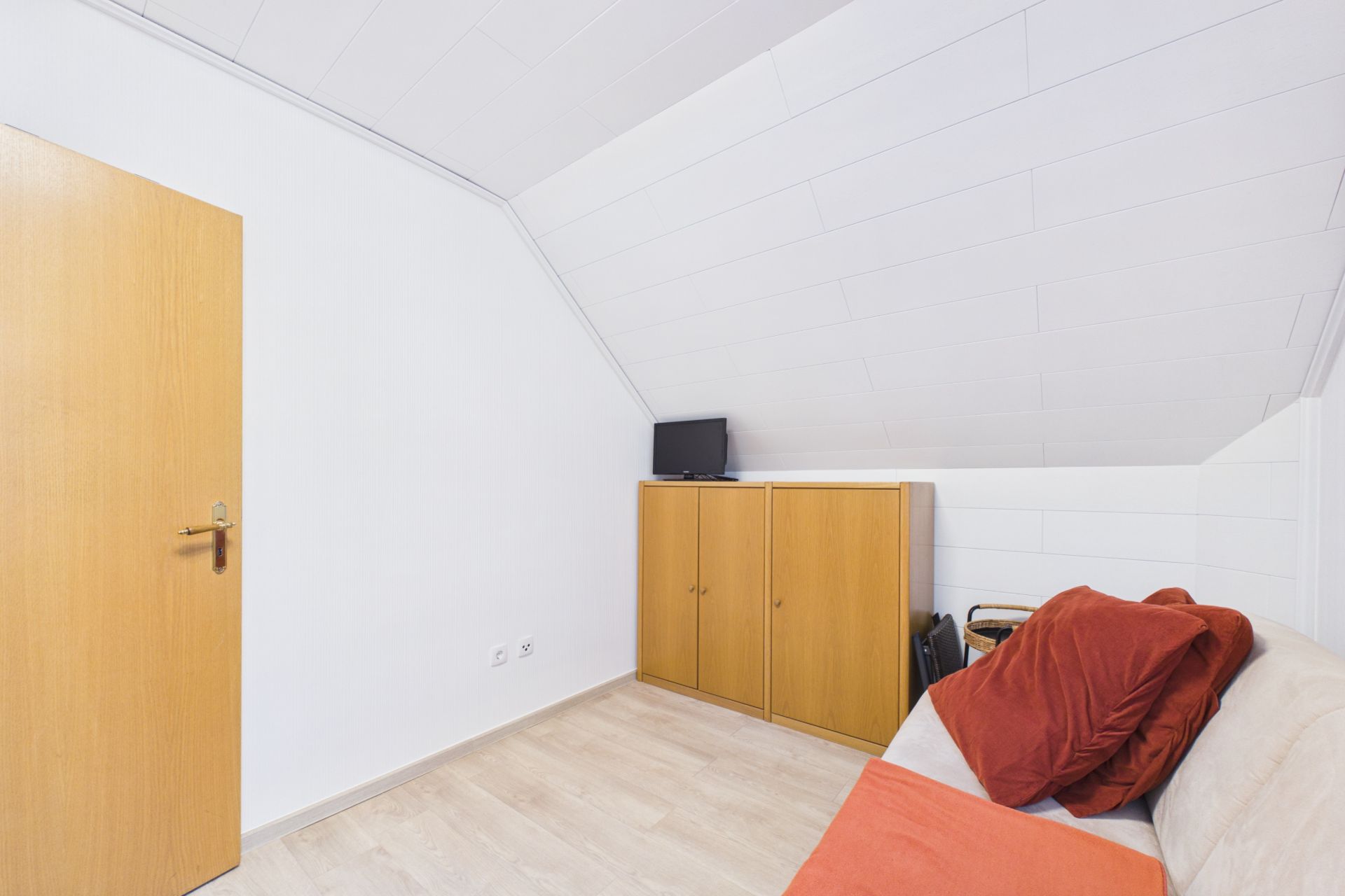 Zimmer mit weißer Schräge, Couch mit roten Kissen, Holzschrank und kleinem Fernseher.