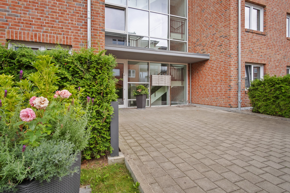 Eingang eines Mehrfamilienhauses aus rotem Backstein mit Blumen und Hecken am Weg.