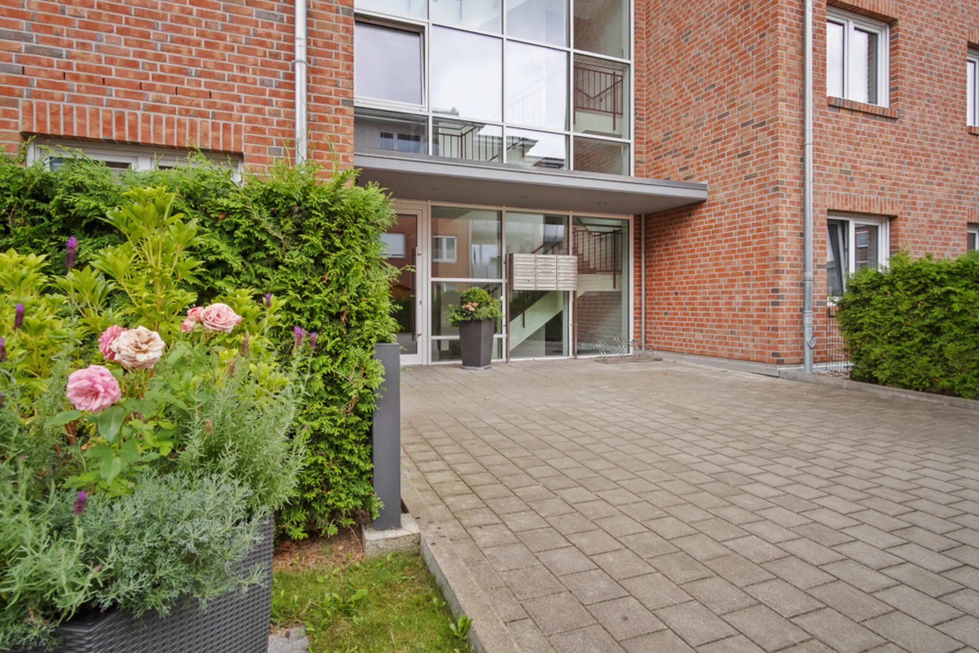 Eingang eines Mehrfamilienhauses aus rotem Backstein mit Blumen und Hecken am Weg.