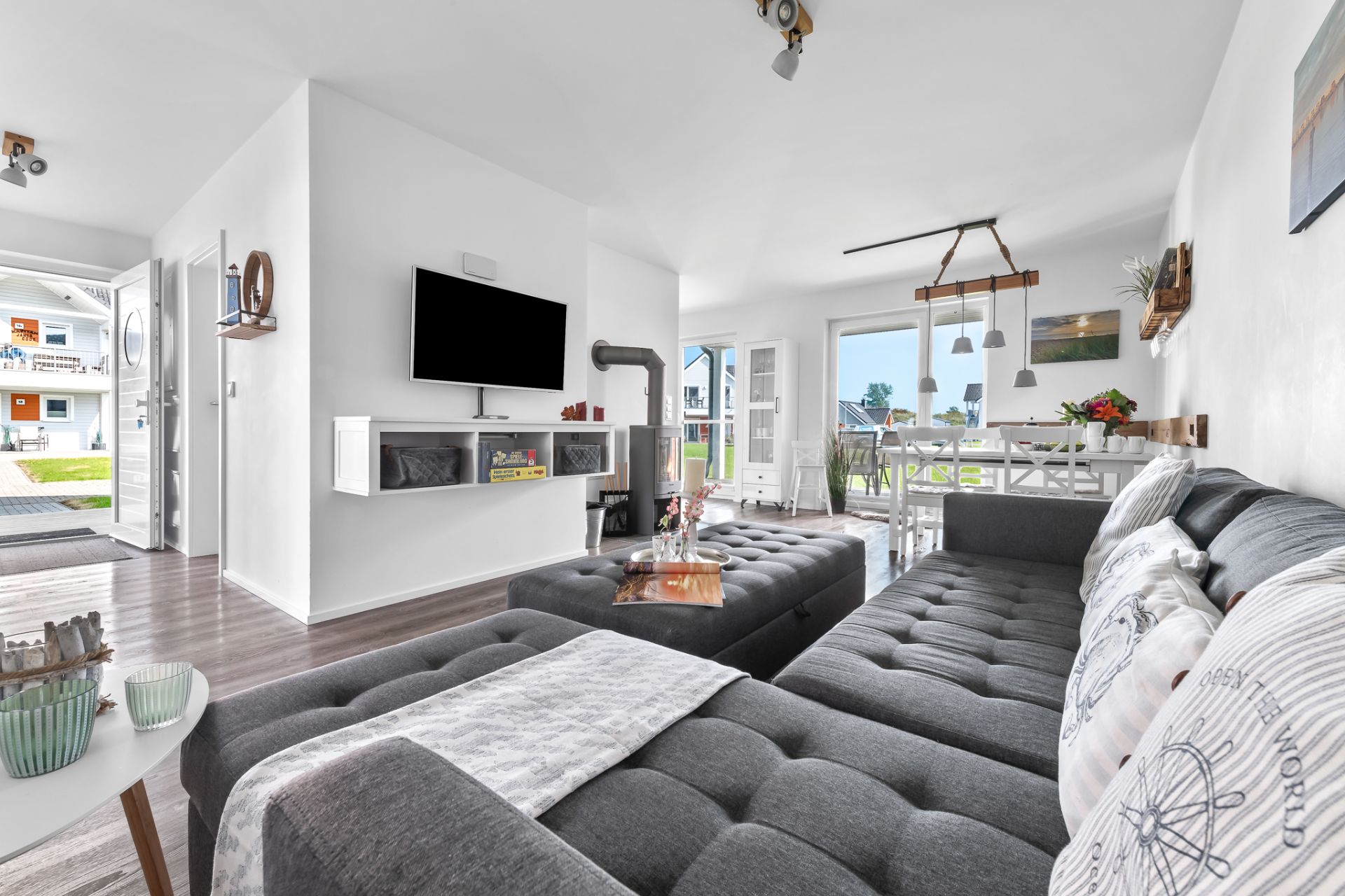 Helles Wohnzimmer mit grauem Sofa, TV an der Wand, Kamin und Esstisch, große Fenster zur Terrasse.