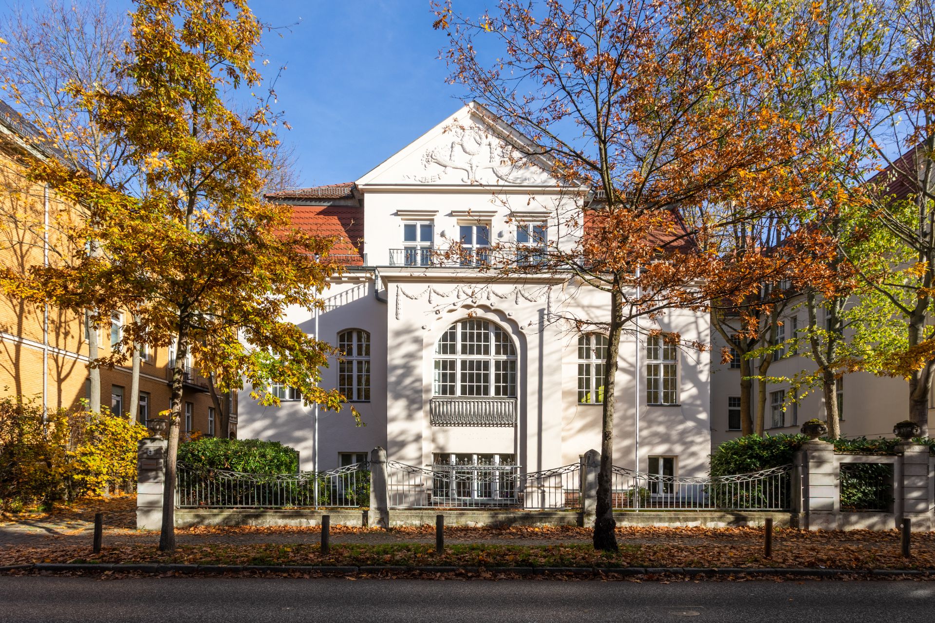 Weiße Wohnvilla mit Ziergiebel, großen Fenstern und umliegenden Bäumen im Herbst.