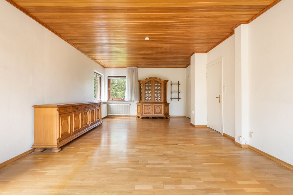 Helles Wohnzimmer mit Holzdecke, Holzboden, Kommode, Vitrine und zwei Fenstern.