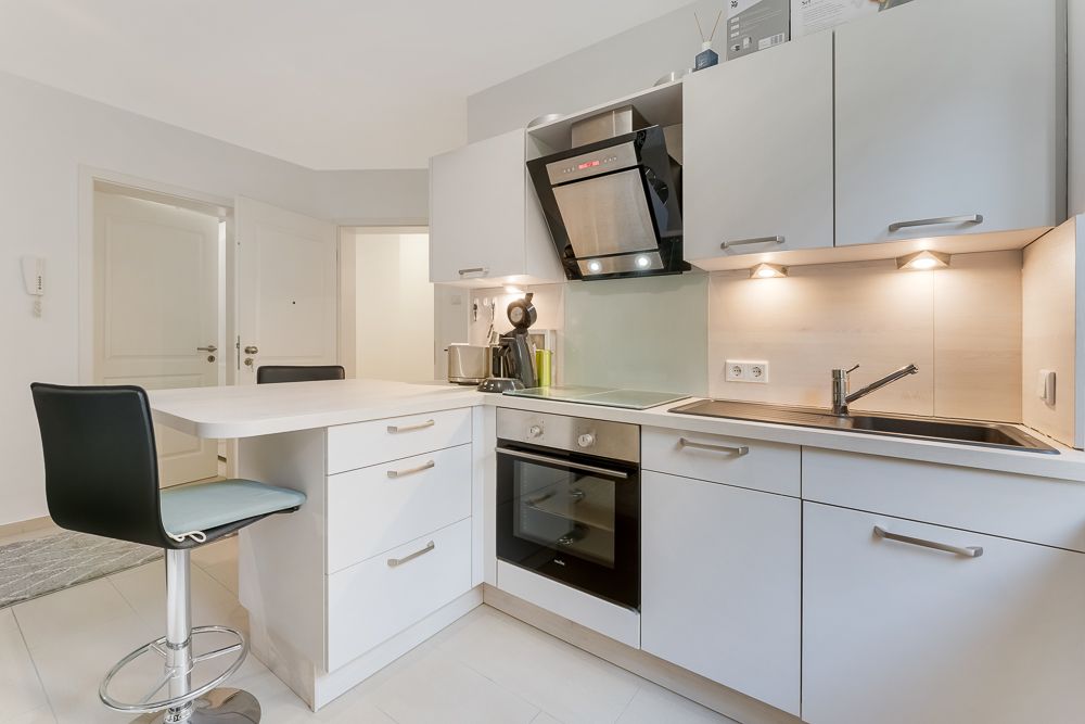 Moderne Einbauküche mit Kochfeld, Backofen, Spüle, Oberschränken und Barhocker.