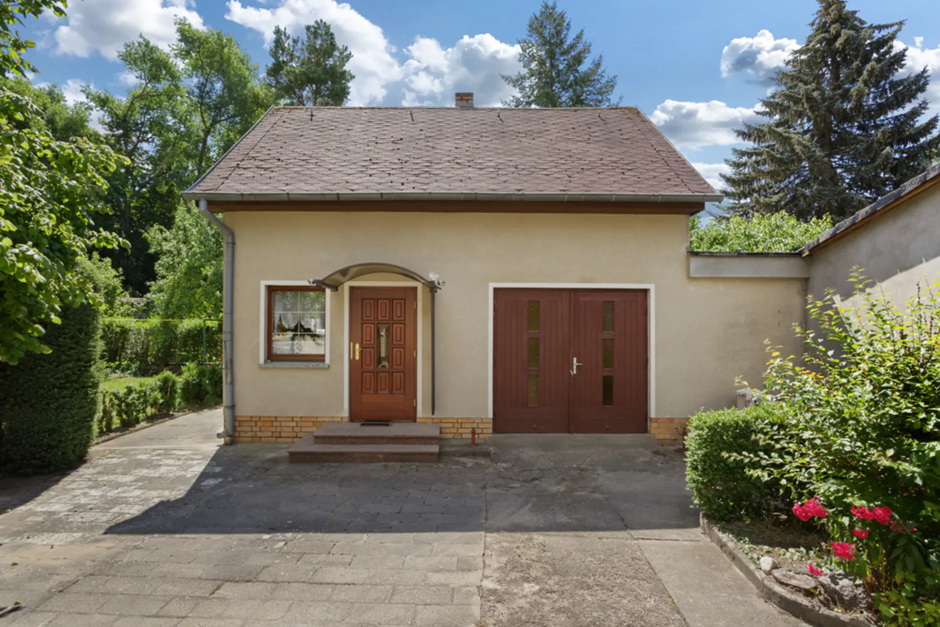 Vorderansicht eines kleinen Hauses mit brauner Tür, Garage und Garten im Hintergrund.