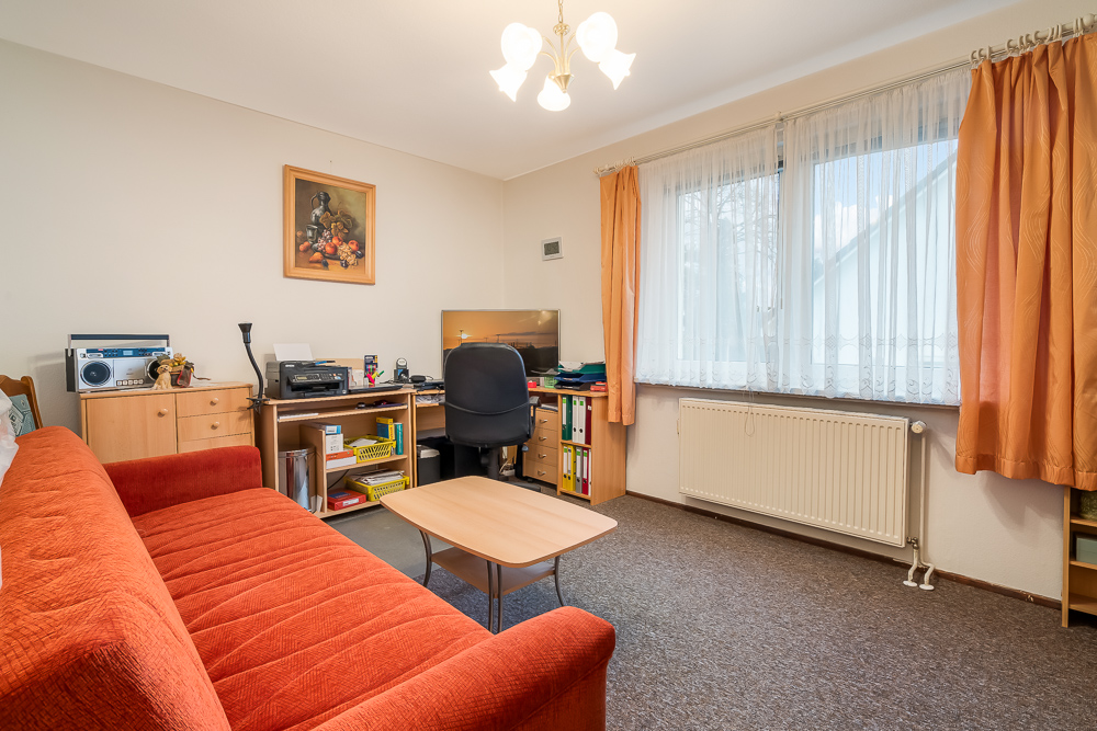 Wohnzimmer mit rotem Sofa, Schreibtisch, Bürostuhl und großem Fenster mit orangefarbenen Vorhängen.