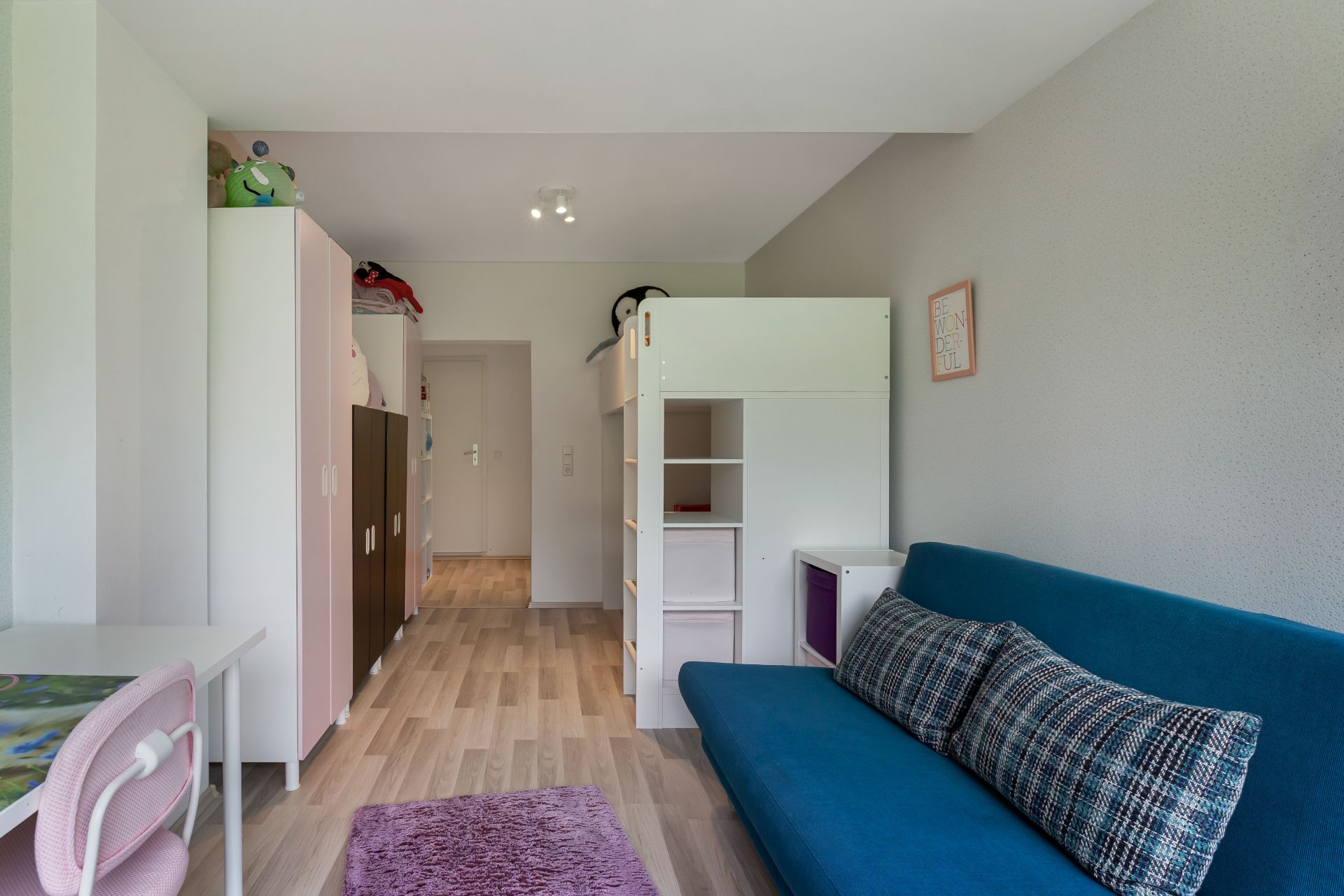 Kinderzimmer mit Hochbett, blauem Sofa, Schreibtisch und mehreren Schränken auf Holzfußboden.
