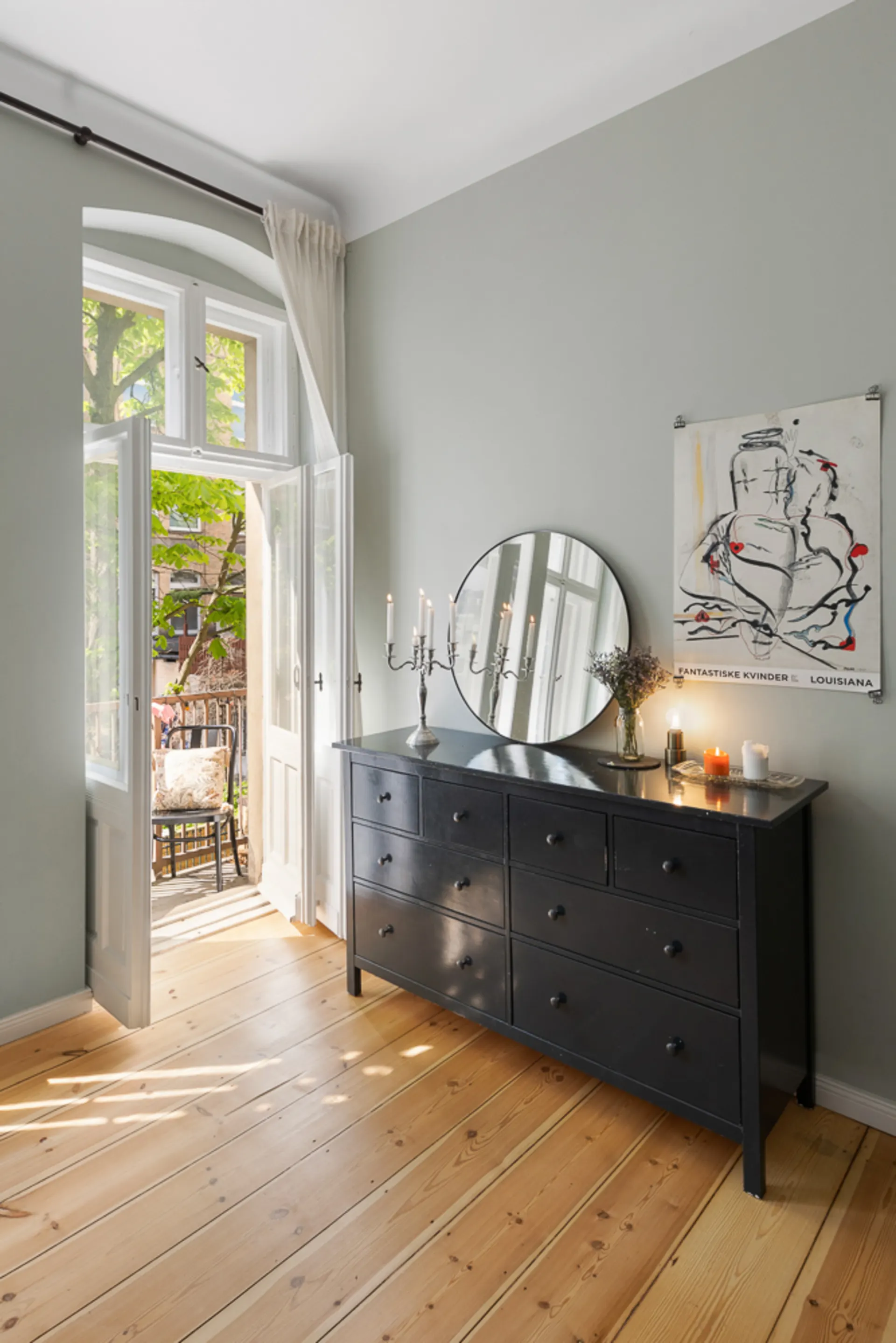 Helles Zimmer mit Holzboden, Kommode, rundem Spiegel und offenem Fenster zur Terrasse.