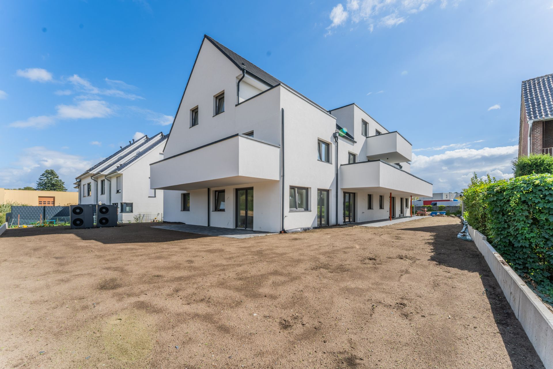 Modernes, weißes Wohnhaus mit Balkonen und großem, unbepflanztem Vorhof bei Sonnenschein.