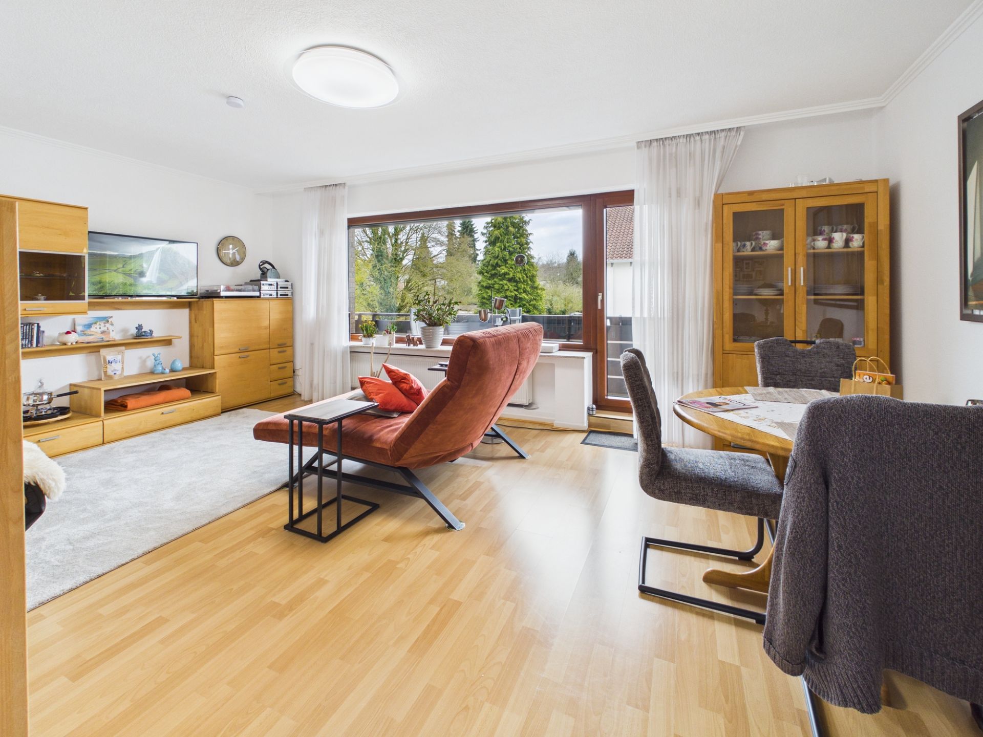 Helles Wohnzimmer mit Holzboden, Sofa, Essbereich, Schrankwand und großen Fenstern mit Blick ins Grüne.