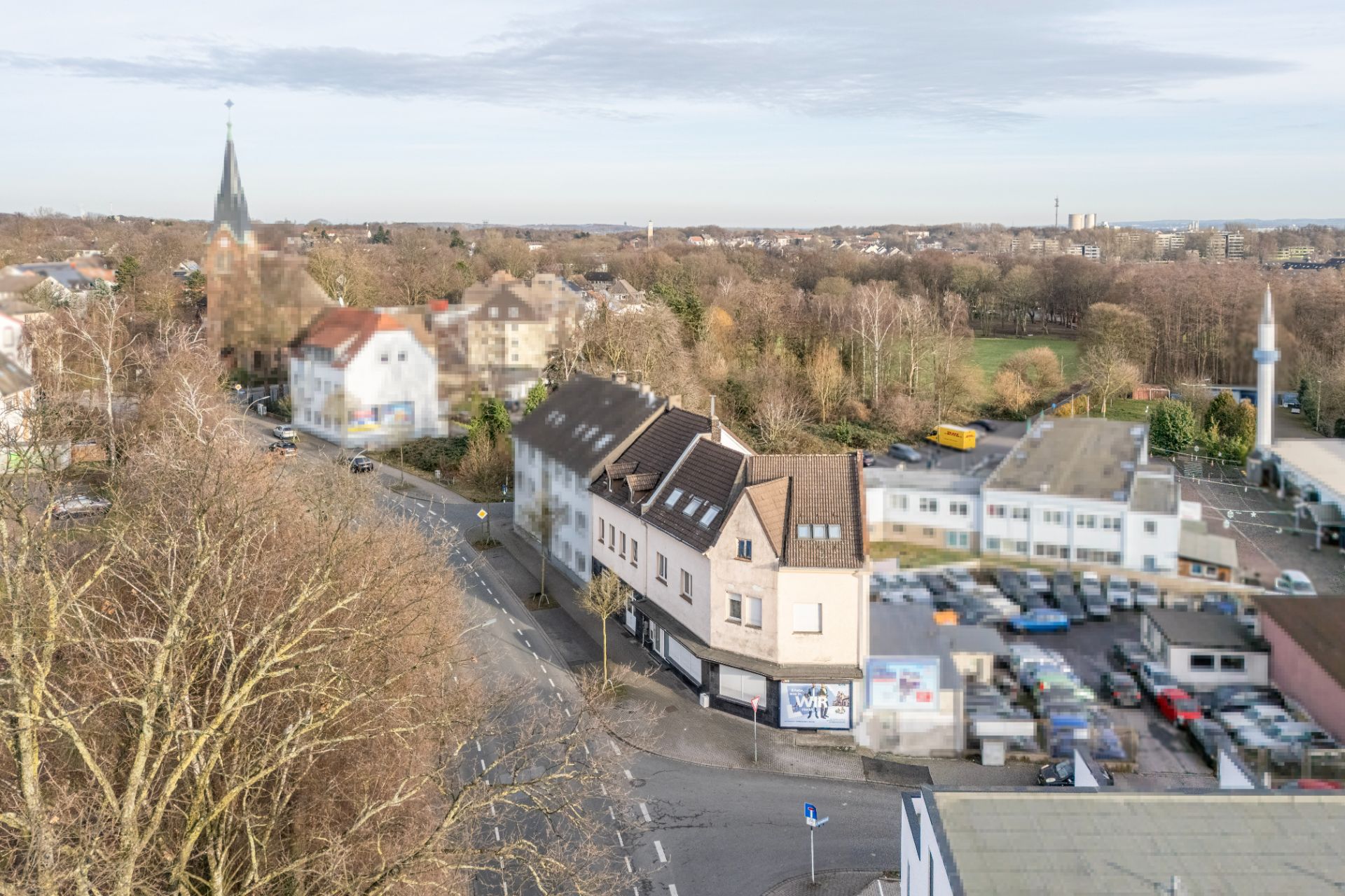 Straßenansicht mit Wohnhäusern, Bäumen und parkenden Autos, im Hintergrund eine Kirche.