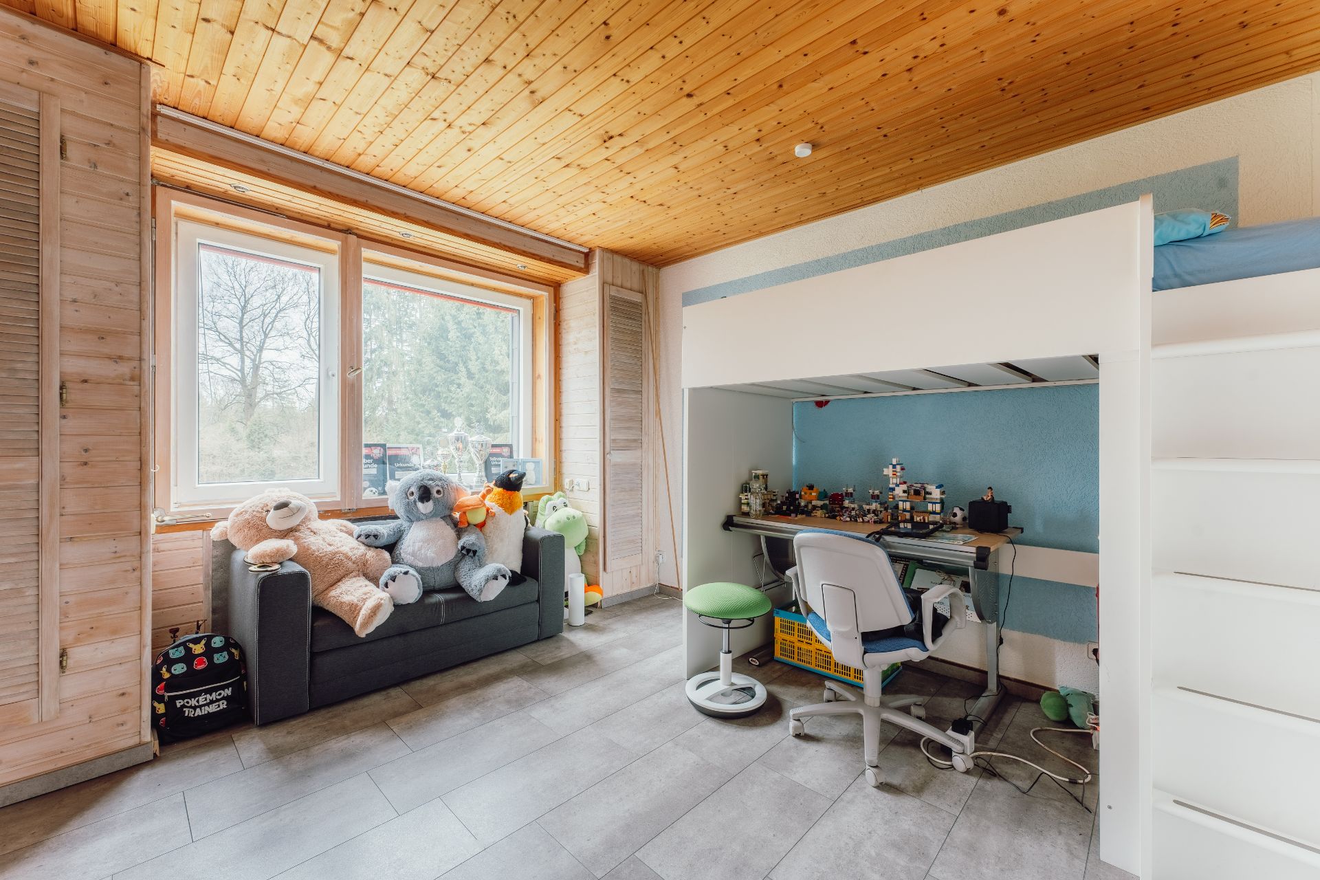 Kinderzimmer mit Hochbett, Schreibtisch, Stuhl, Sofa mit Stofftieren und großem Fenster.