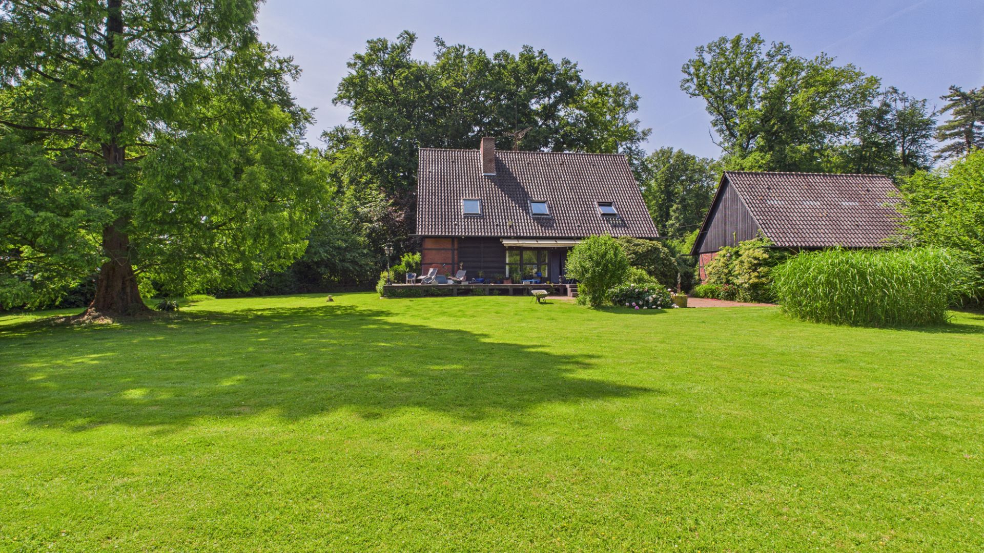 Haus mit Satteldach und Terrasse, großer grüner Garten, Bäume im Hintergrund.