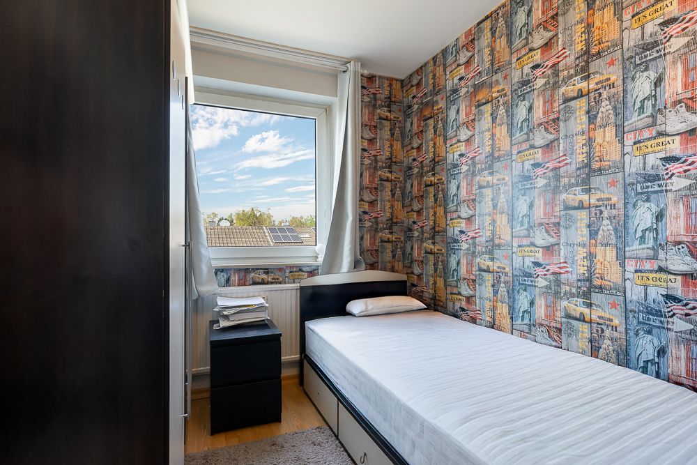 Kleines Schlafzimmer mit Einzelbett, Nachttisch und bunter Tapete, Fenster mit Blick nach draußen.