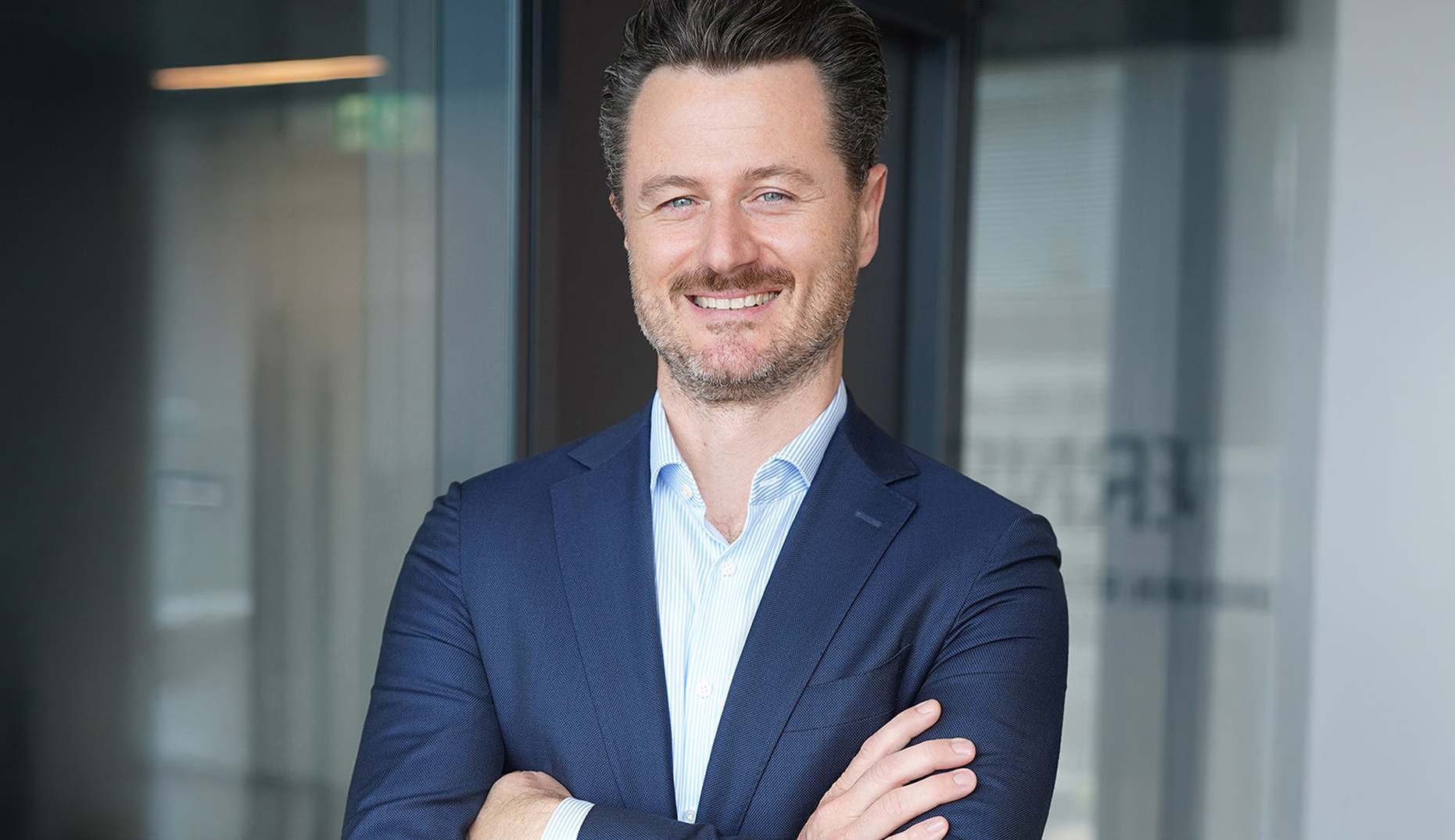 Ein Mann in blauem Anzug steht mit verschränkten Armen in einem modernen Innenraum.