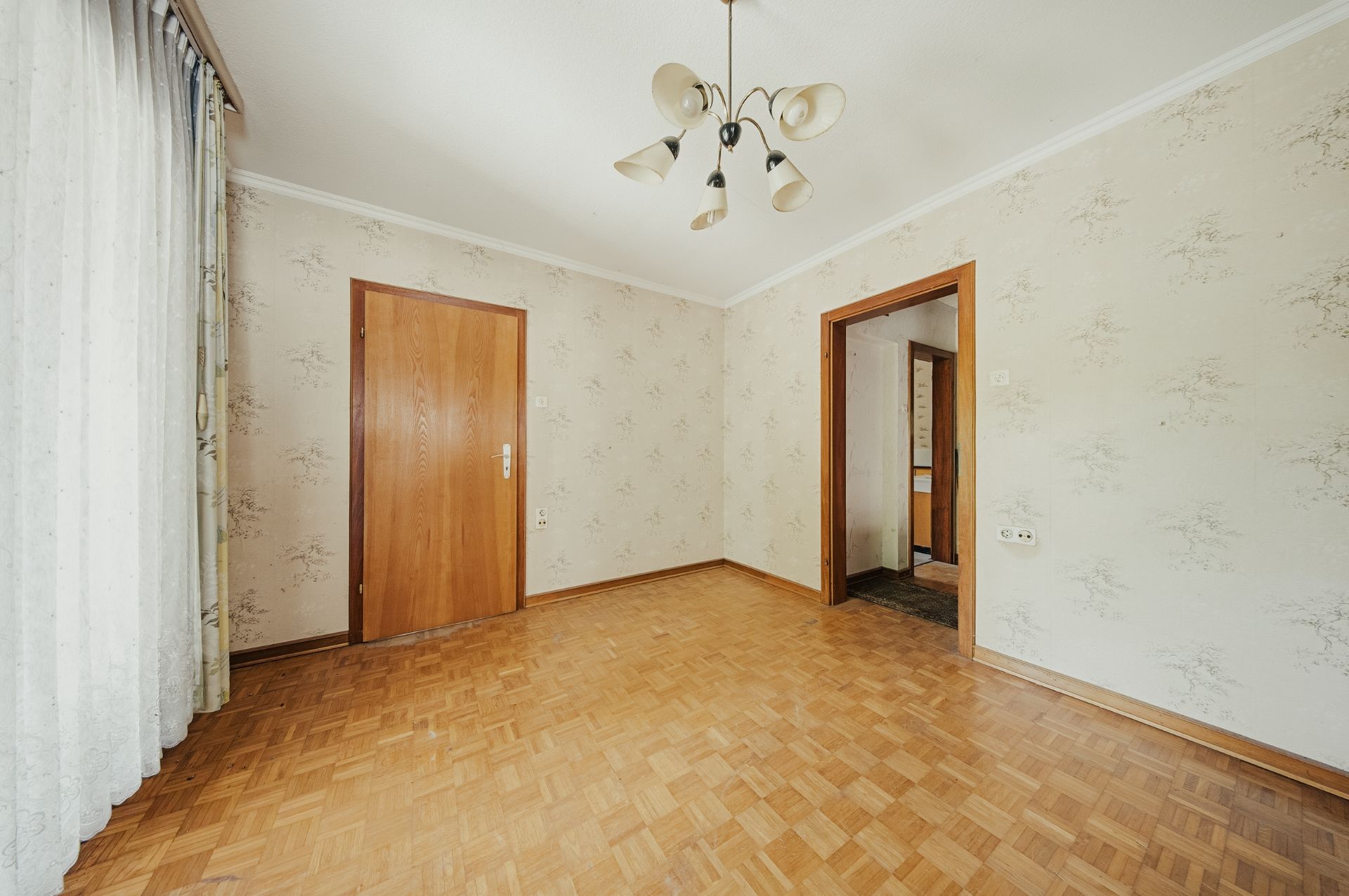 Leeres Zimmer mit Parkettboden, Tapeten, Holztür und großem Fenster mit Vorhang.