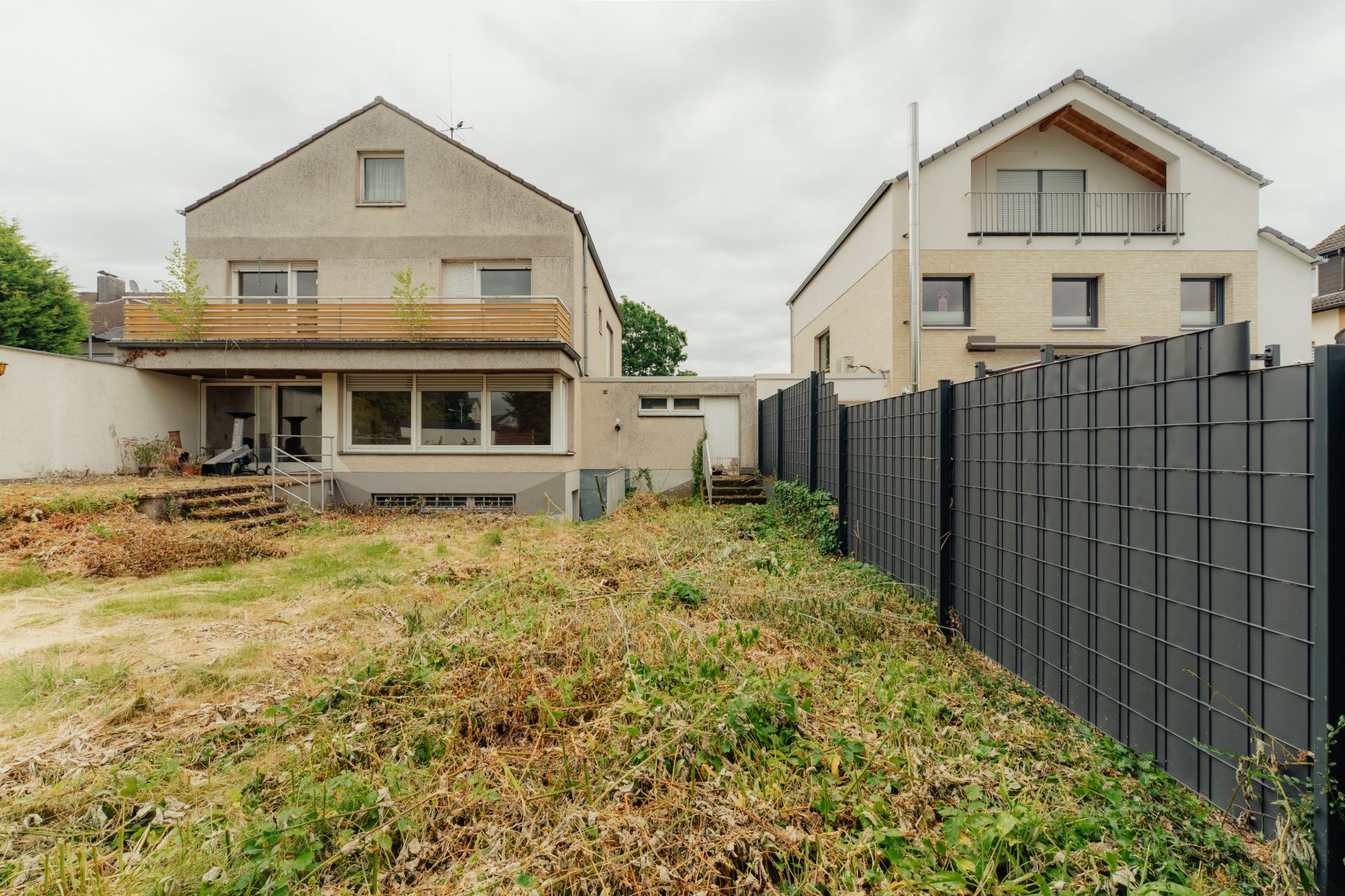 Zwei Wohnhäuser mit Garten und Zaun, teilweise bewachsener Boden, Terrasse und Balkon sichtbar.