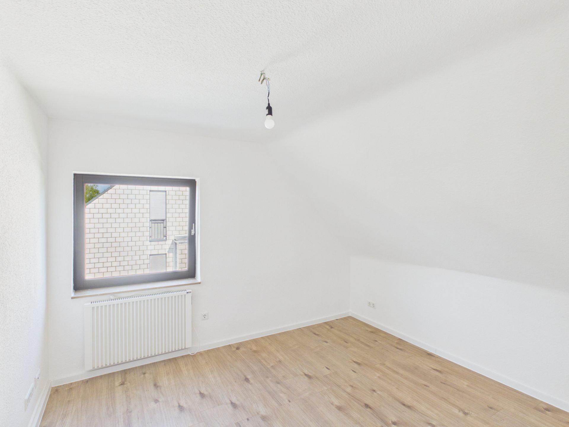 Leeres, helles Zimmer mit weißer Wand, Fenster, Heizkörper und Holzfußboden.