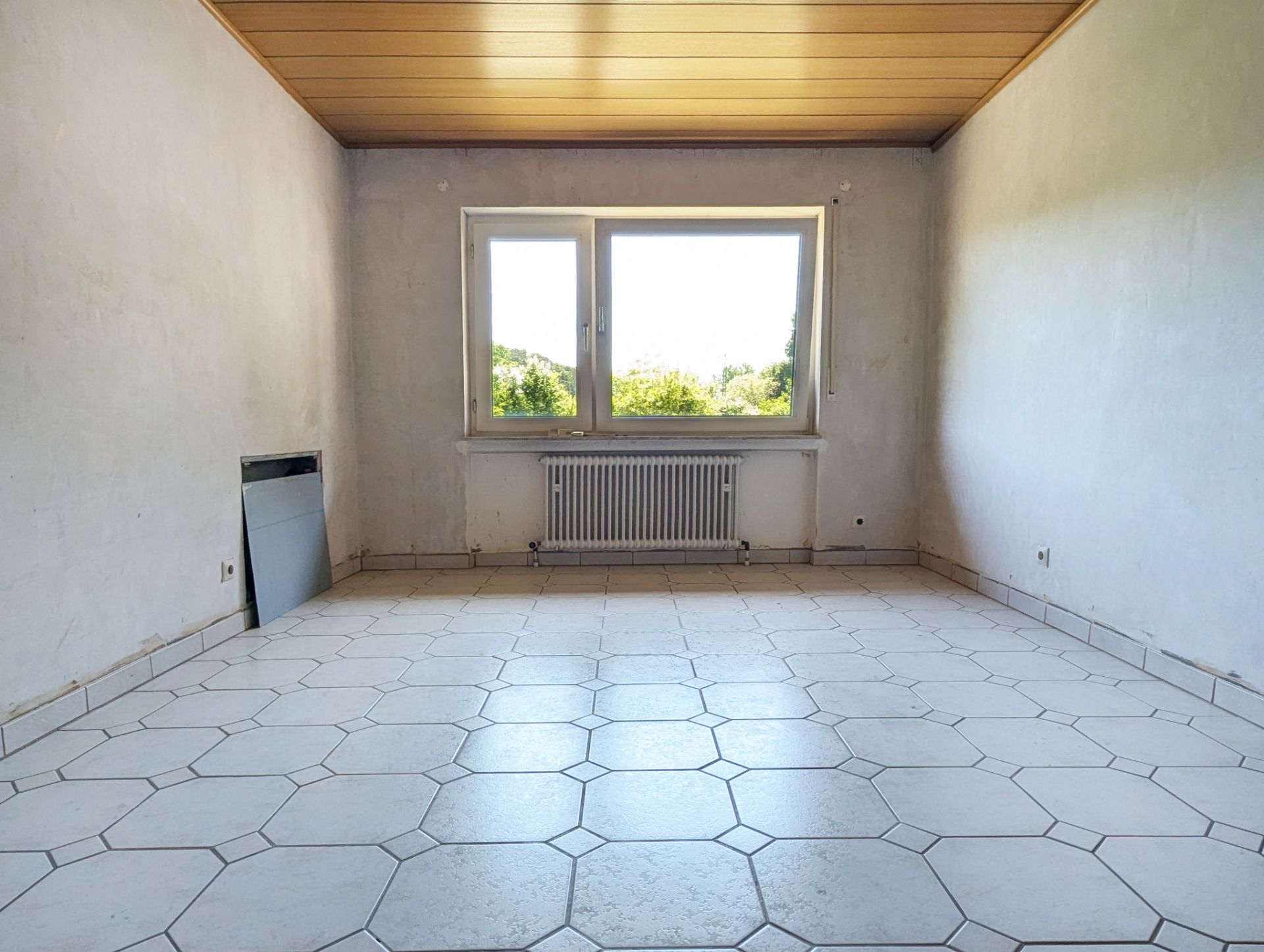 Leeres Zimmer mit weißen Fliesen, großem Fenster und Holzdecke.