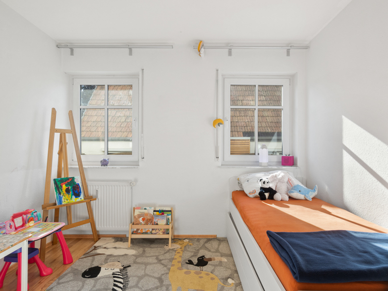 Helles Kinderzimmer mit zwei Fenstern, Bett, Spielzeug, Tisch, Bücherregal und bunter Spielmatte.