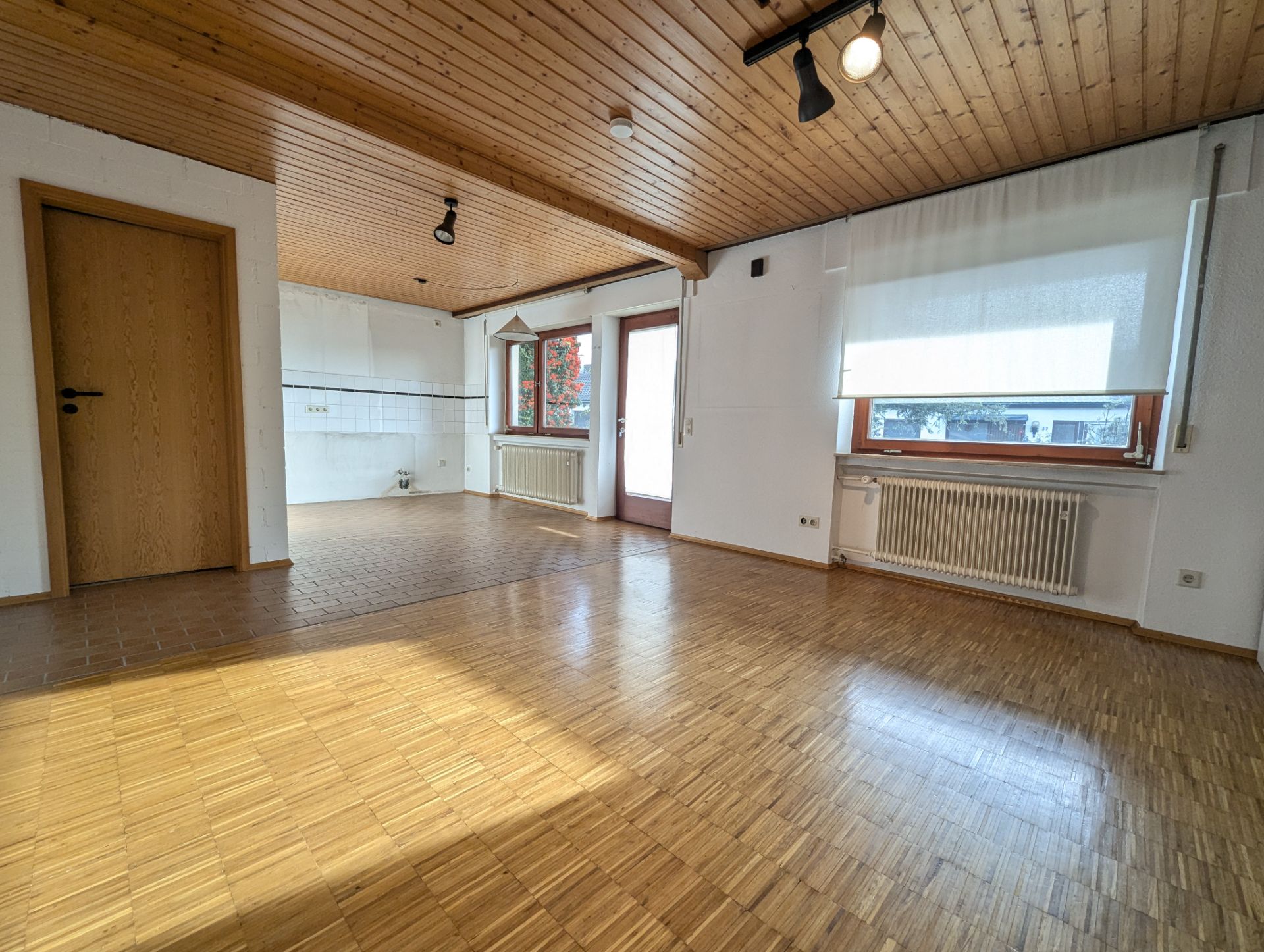 Helles Wohnzimmer mit Holzboden und Decke, angrenzender offener Küche, großen Fenstern und Tür zum Balkon.