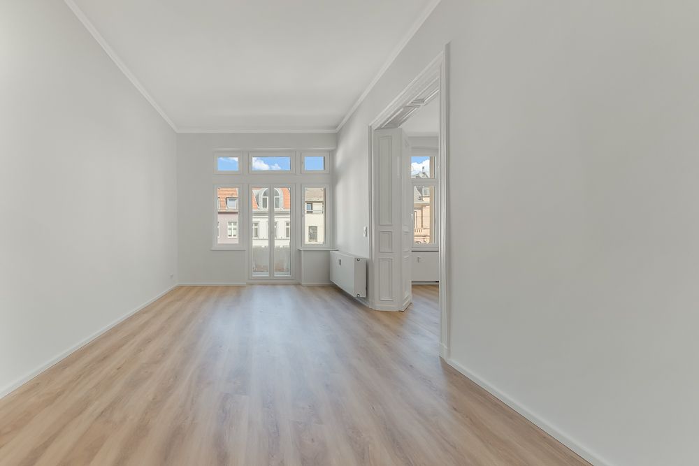 Heller, leerer Raum mit Holzfußboden und großen Fenstern, Blick auf Nachbarhäuser.