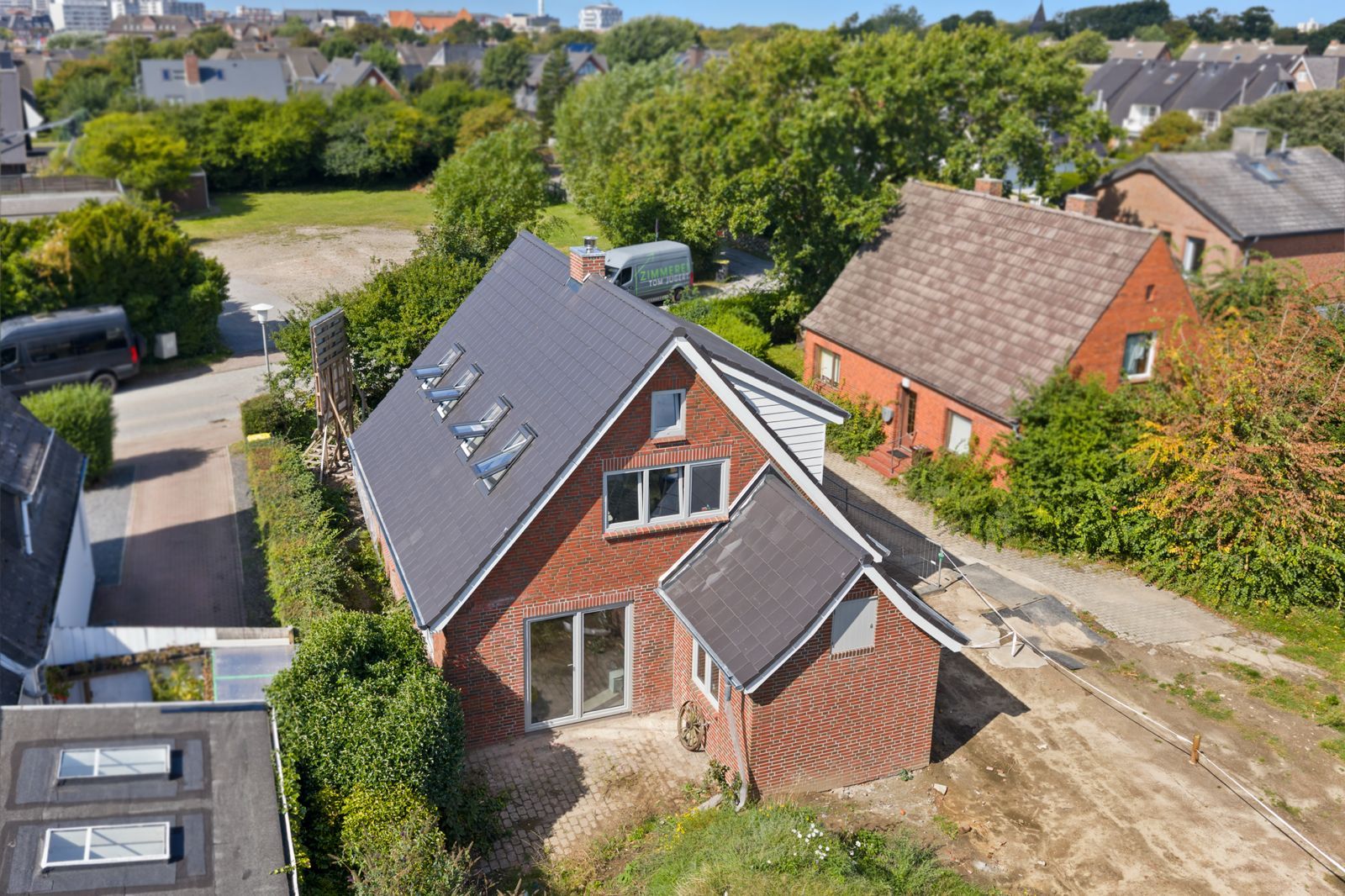 Backstein-Einfamilienhaus mit großem Dach und mehreren Dachfenstern, umgeben von Garten und Nachbarhäusern.