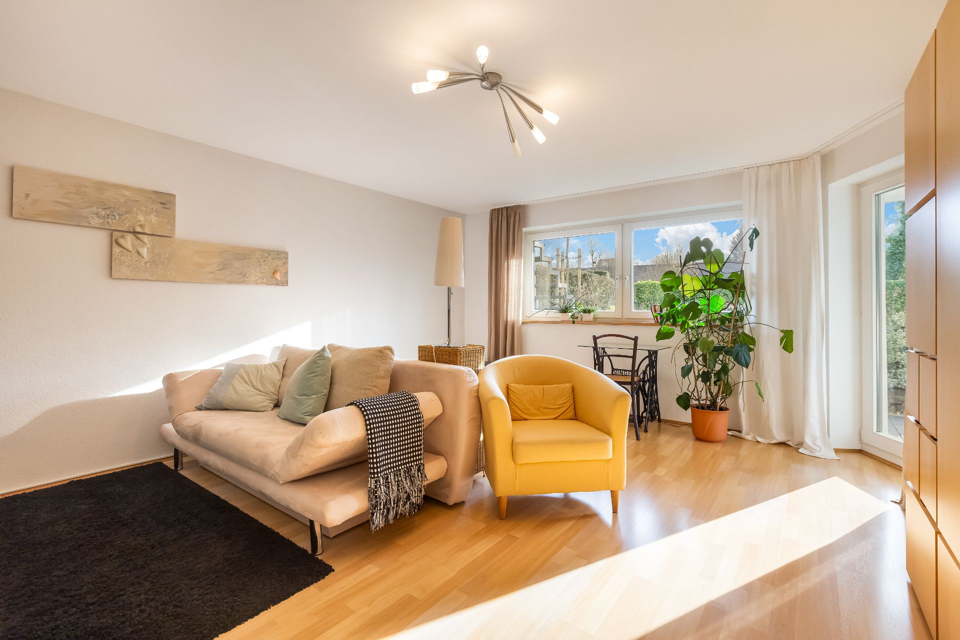 Helles Wohnzimmer mit Sofa, gelbem Sessel, Esstisch, großer Pflanze und großen Fenstern.